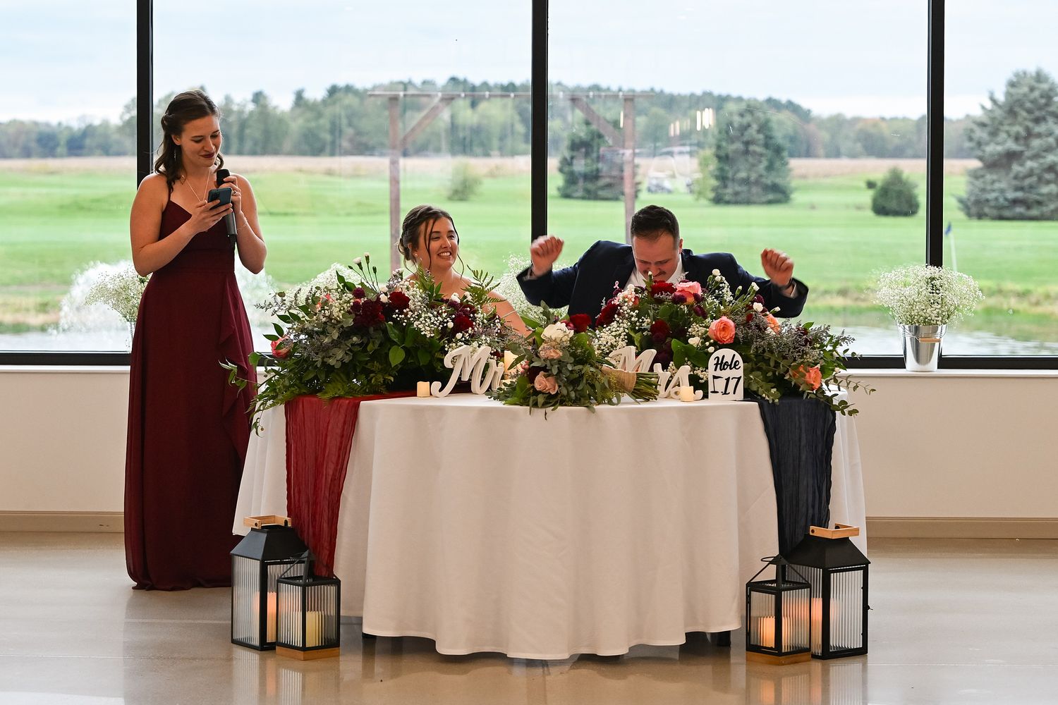 Maid of honor gives a toast speech at wedding reception while bride and groom sit at sweetheart table.