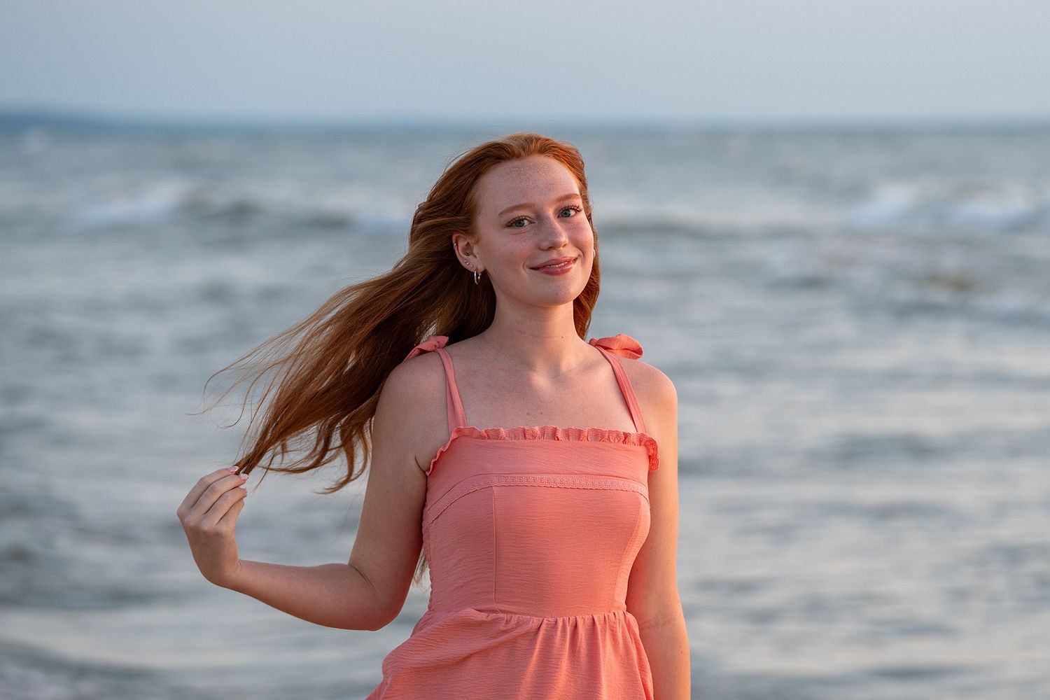 Young redhead woman in pink sundress smiling at beach with ocean waves in background on a breezy day.
