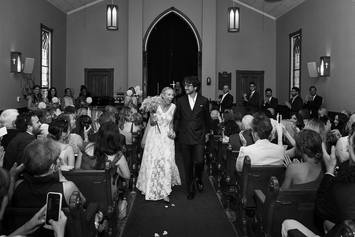 Black and white photo of a joyful couple walking down the aisle after their wedding ceremony in a chapel.