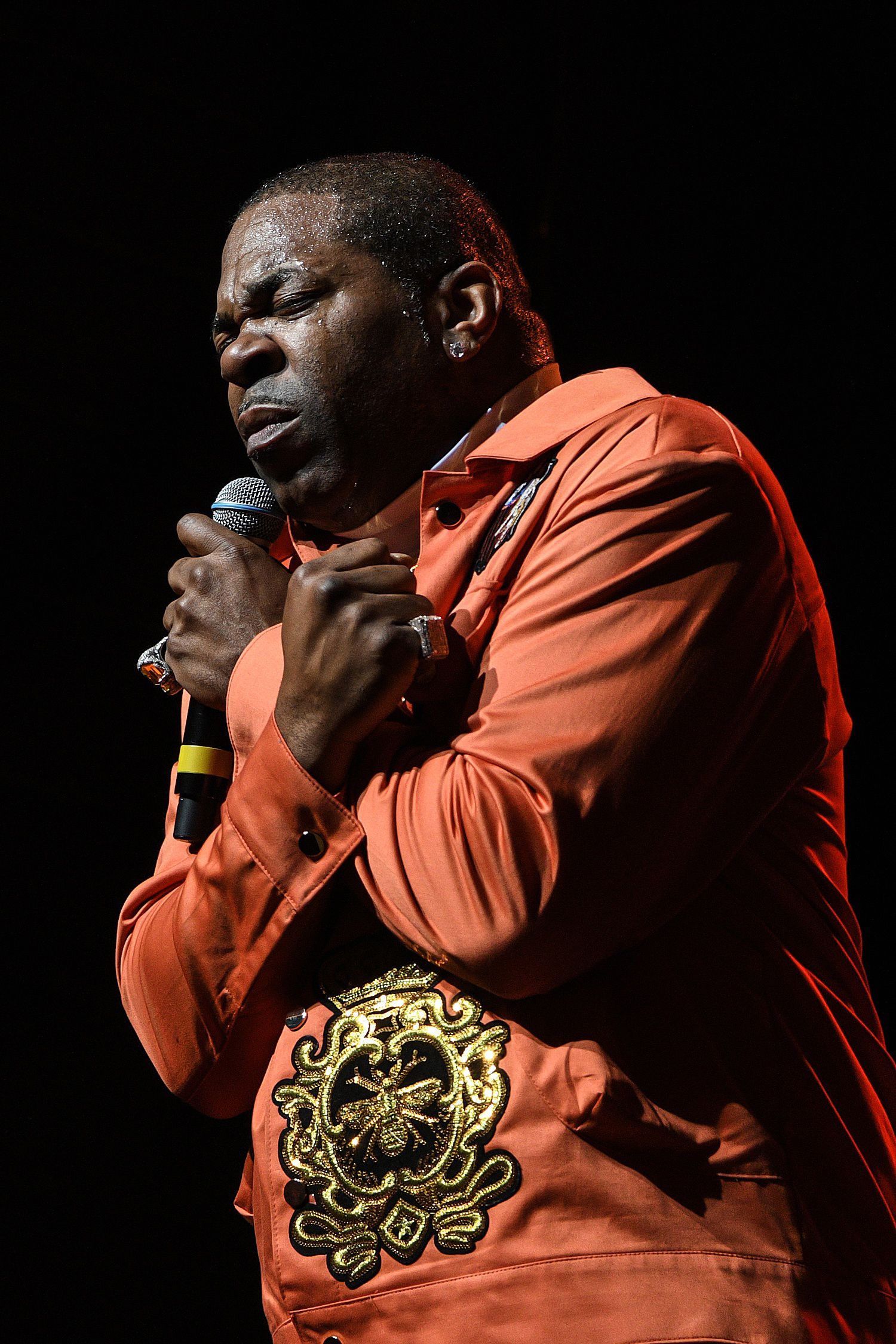Performer in orange jacket singing emotionally into microphone on dark stage.