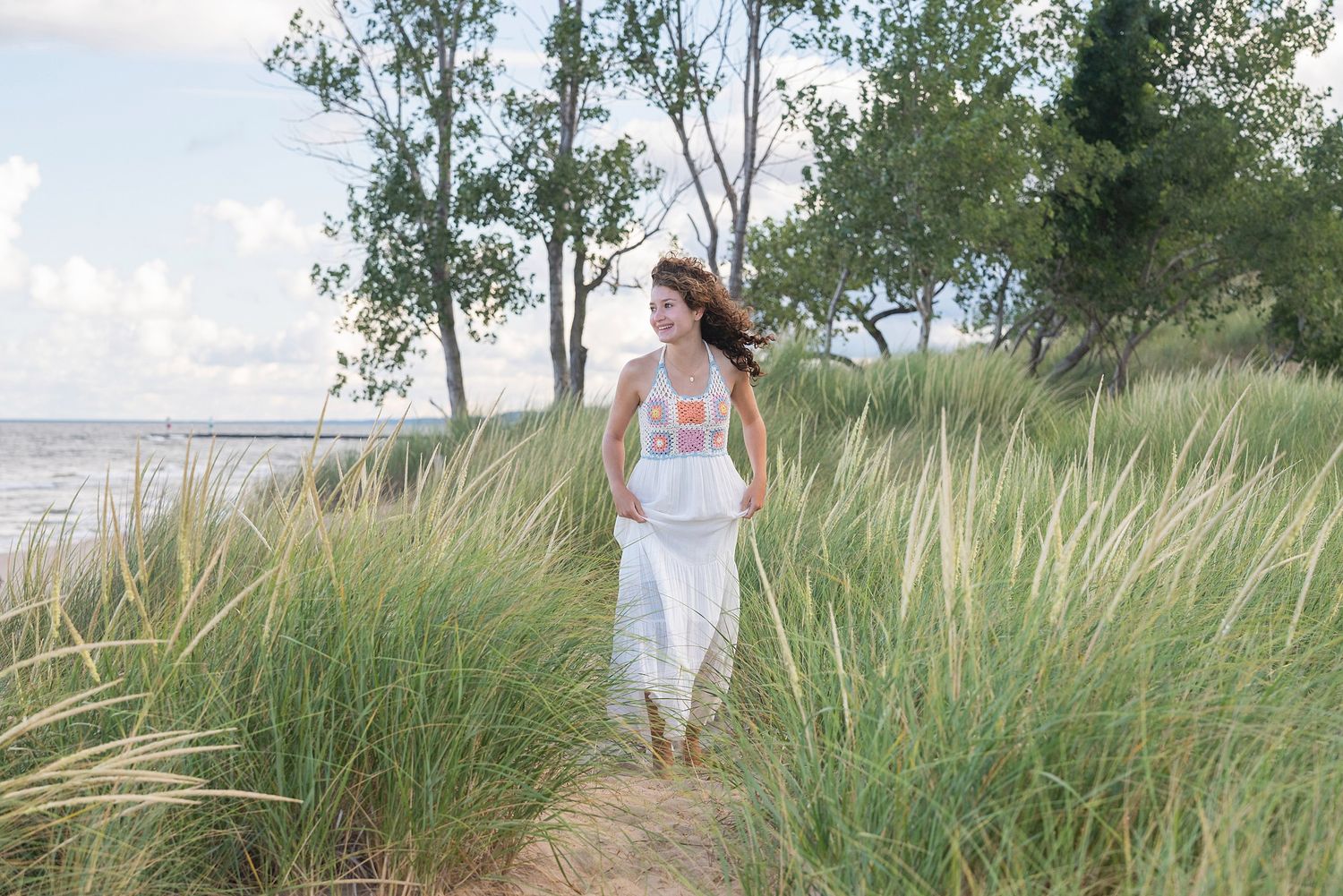 A person in a flowing white dress stands gracefully among tall beach grass on a summer day.