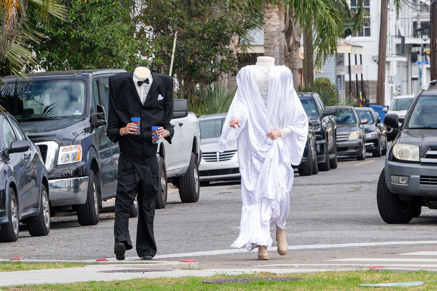 Two figures in ghostly costumes walk down a street lined with parked cars.