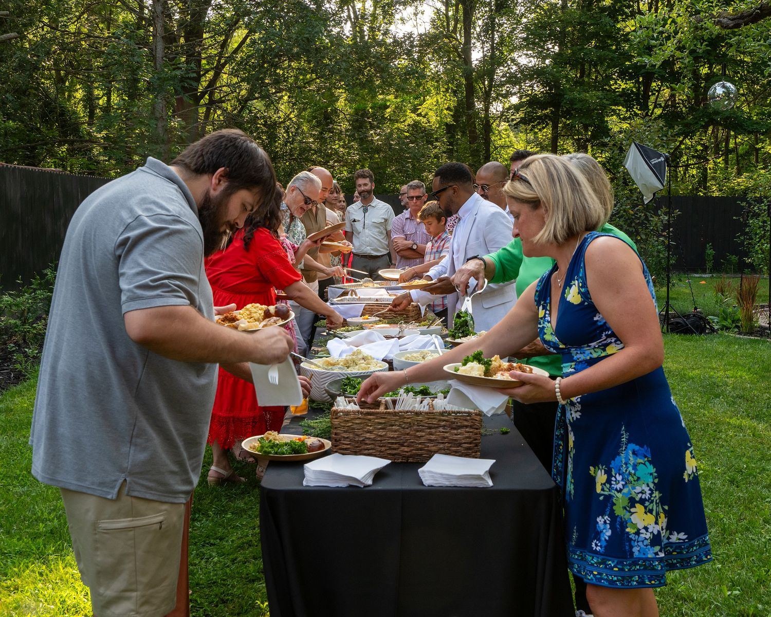 People gather around outdoor tables for a summer barbecue feast in a lush green backyard setting.