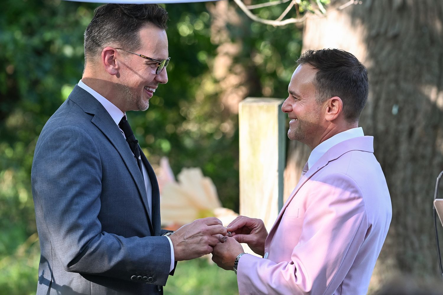 Two people in formal attire share a tender moment during a wedding ceremony in an outdoor garden setting.