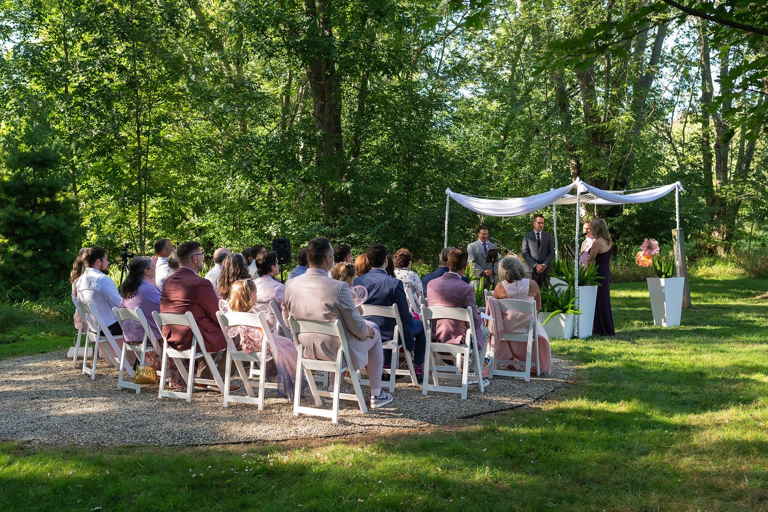 An outdoor wedding ceremony in a wooded setting with white chairs and a decorative canopy on a sunny summer day.