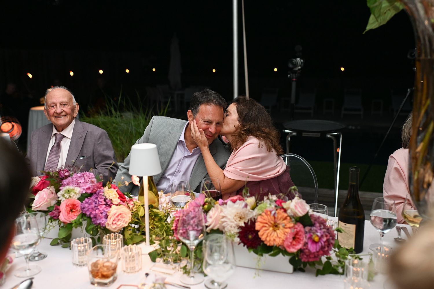 Guests share intimate moments at an elegant outdoor dinner reception with pink floral centerpieces and ambient lighting.