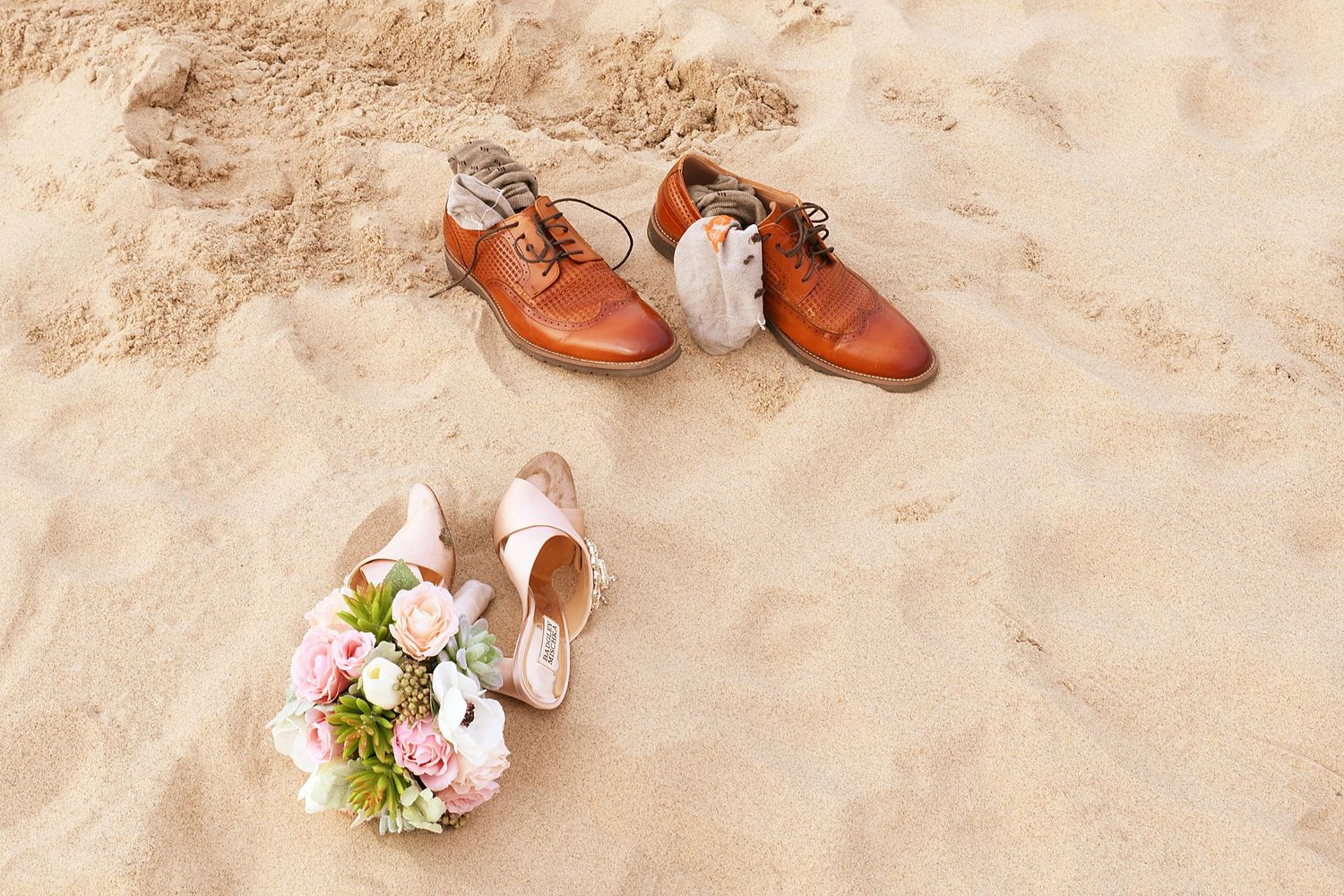 Brown leather wedding shoes placed on sandy beach with floral bouquet.