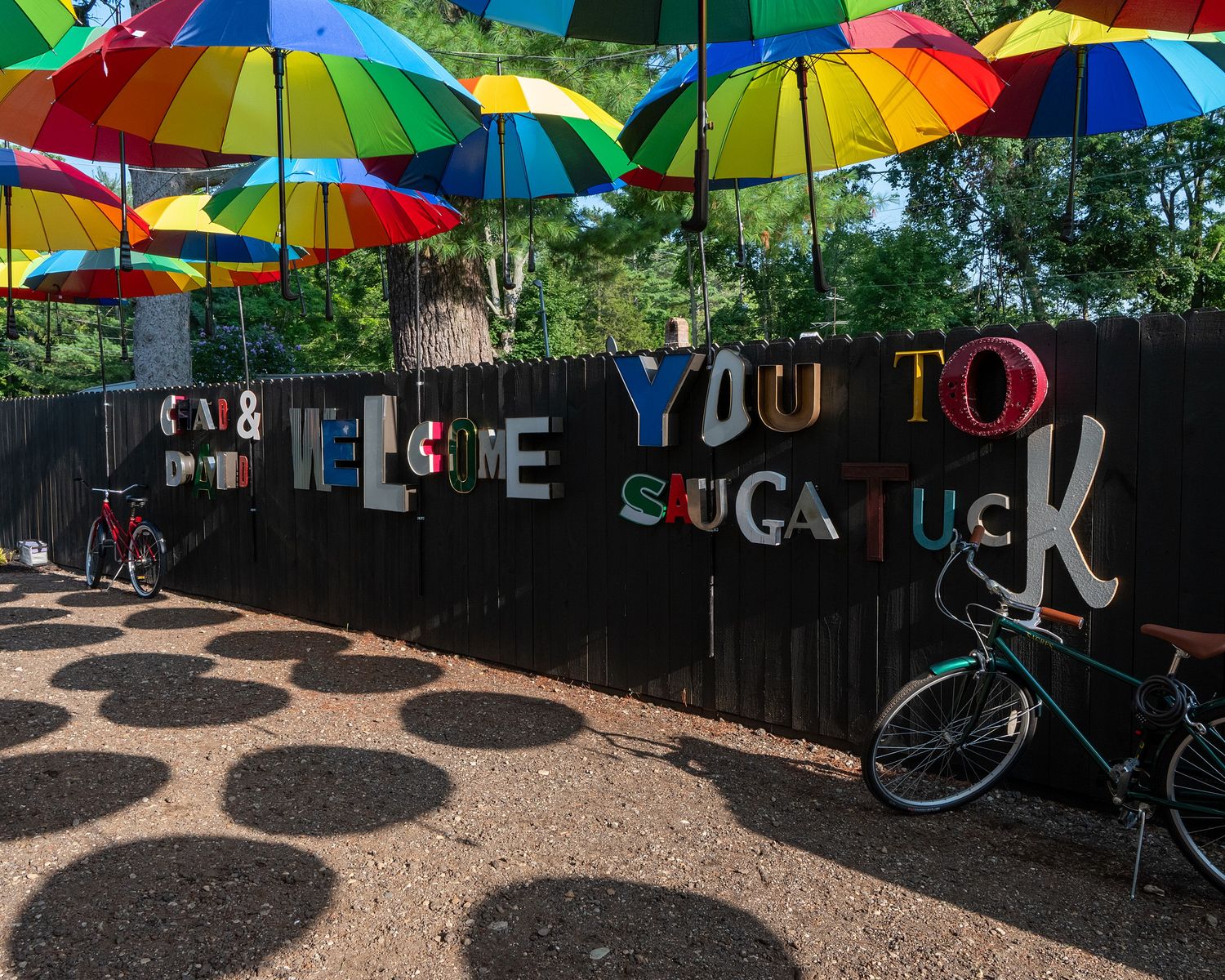 Colorful umbrellas hanging over a garden path create a vibrant canopy against a black fence with decorative text.