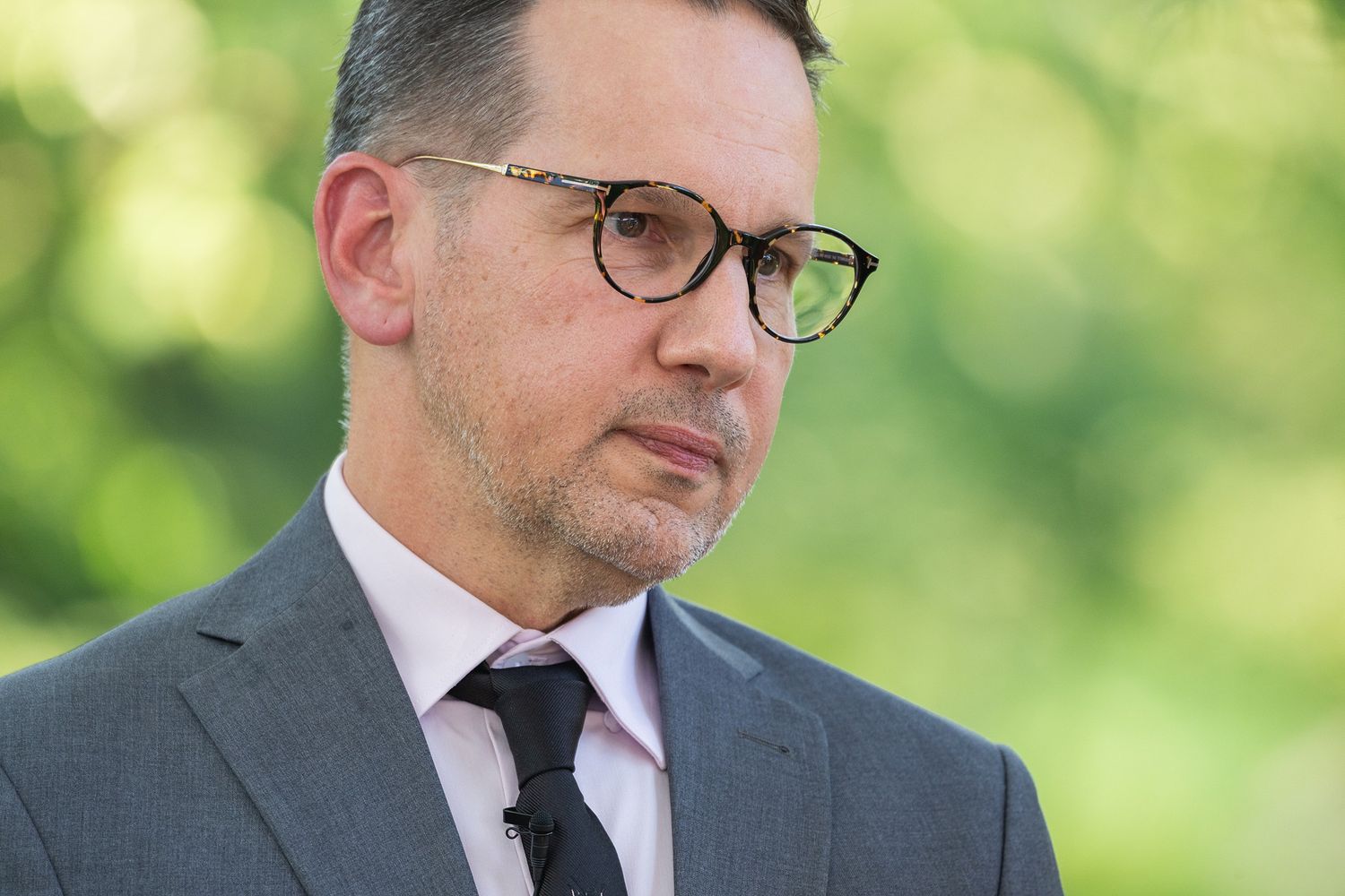 Professional wearing glasses and a dark suit gazes thoughtfully to the side against a blurred green background.