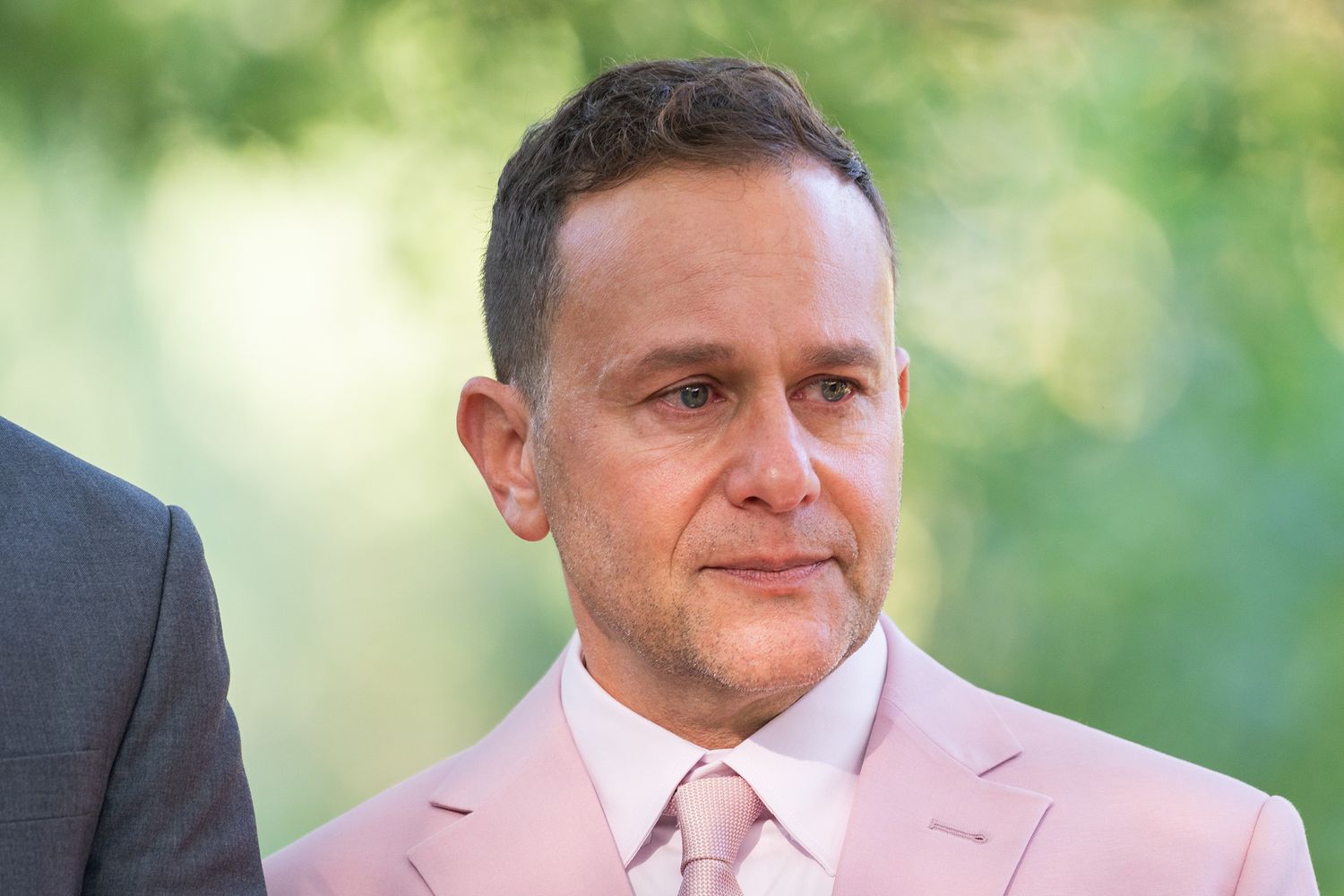 A person wearing a light pink suit and tie stands outdoors with greenery in the background.