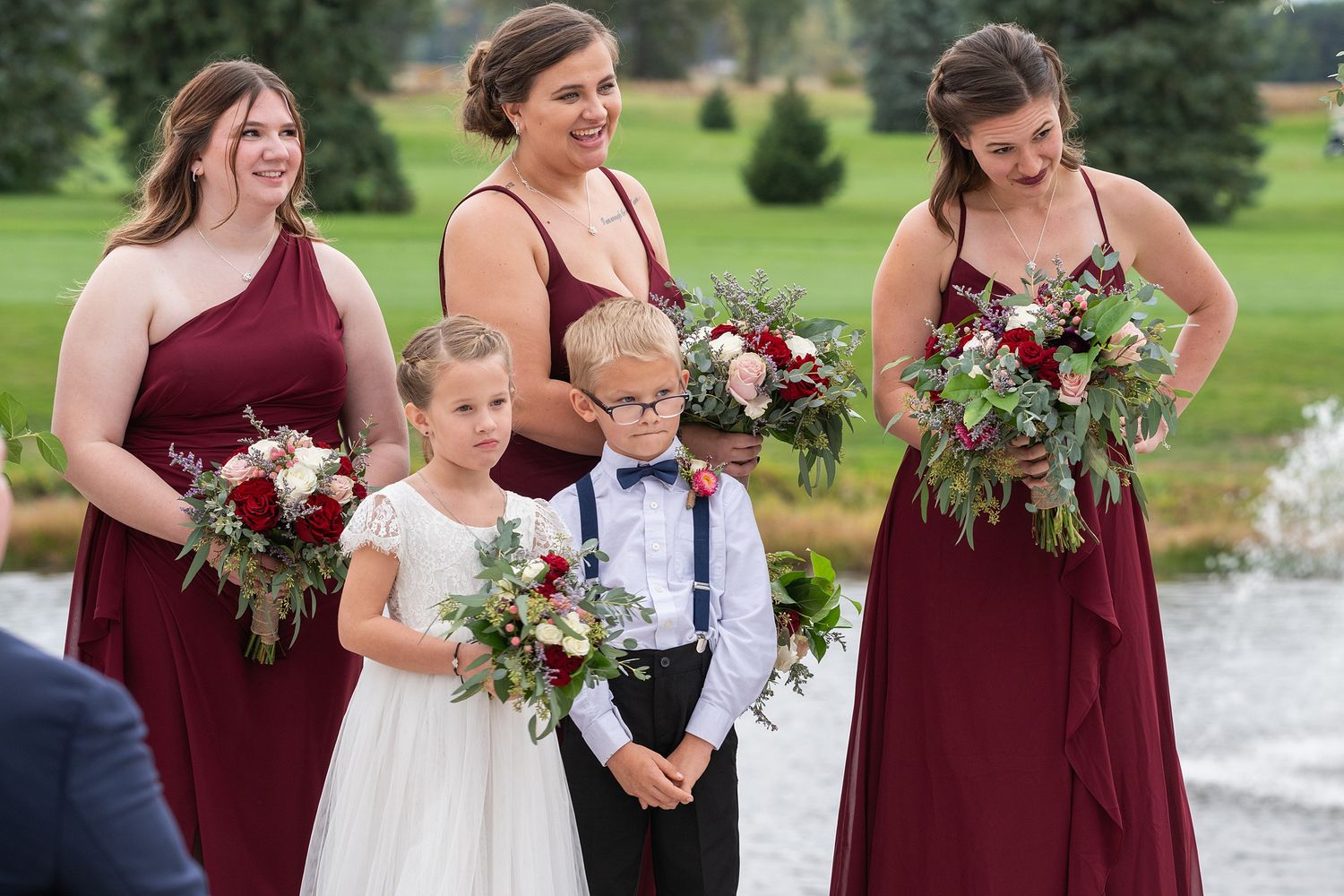 Three bridesmaids in burgundy gowns hold red bouquets alongside flower girl and ring bearer.
