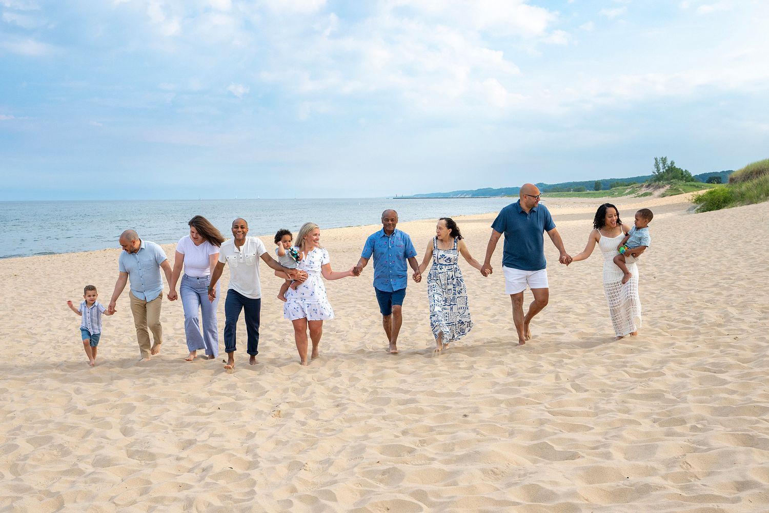 Multi-generational family holding hands walking together on a sunny sandy beach with calm blue ocean waters in the background.