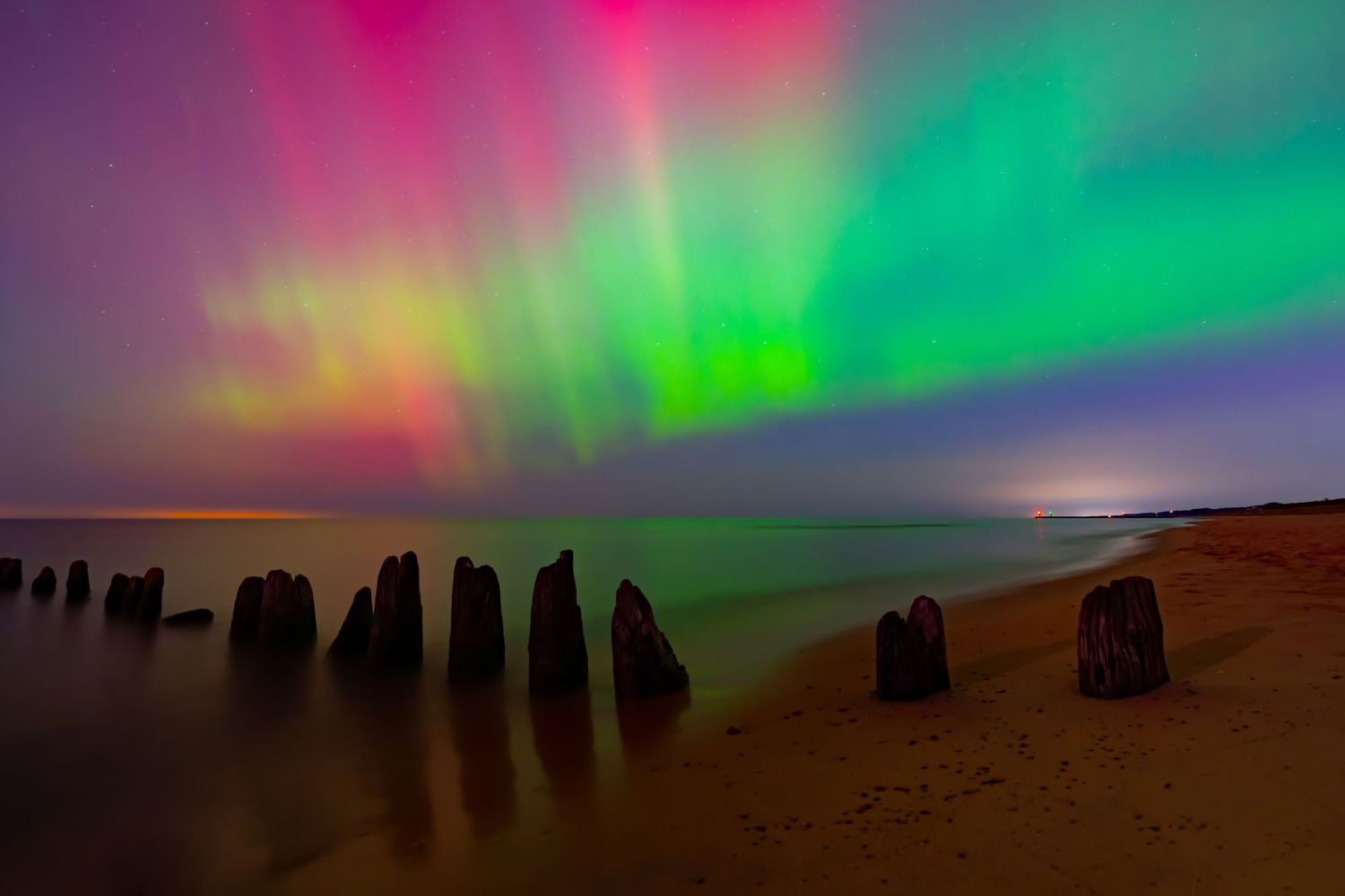 Vibrant aurora borealis illuminates a colorful night sky over a calm beach with weathered wooden posts.