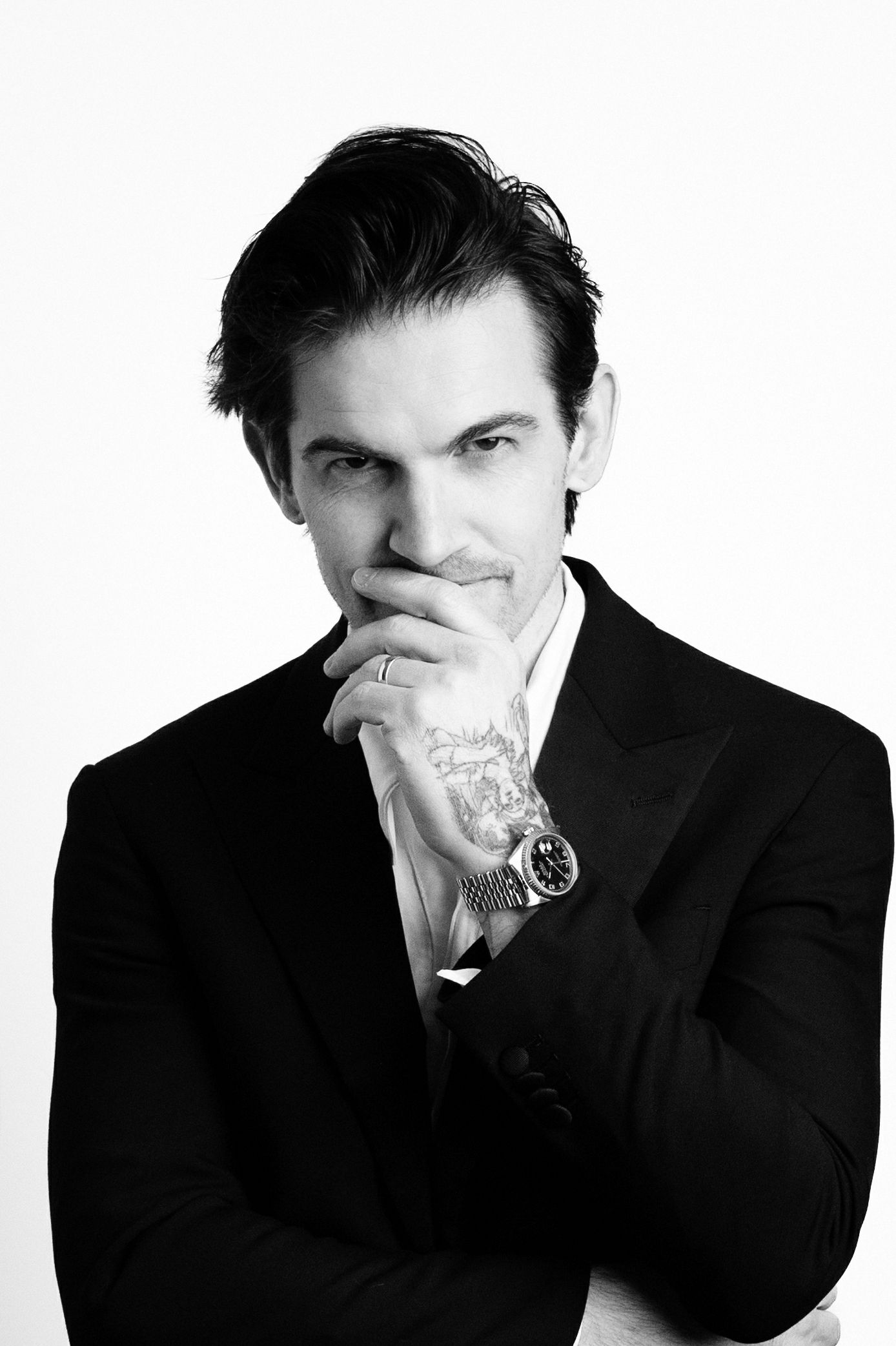 Black and white portrait of a man in a dark suit covering his mouth with a tattooed hand, wearing a wristwatch and ring.