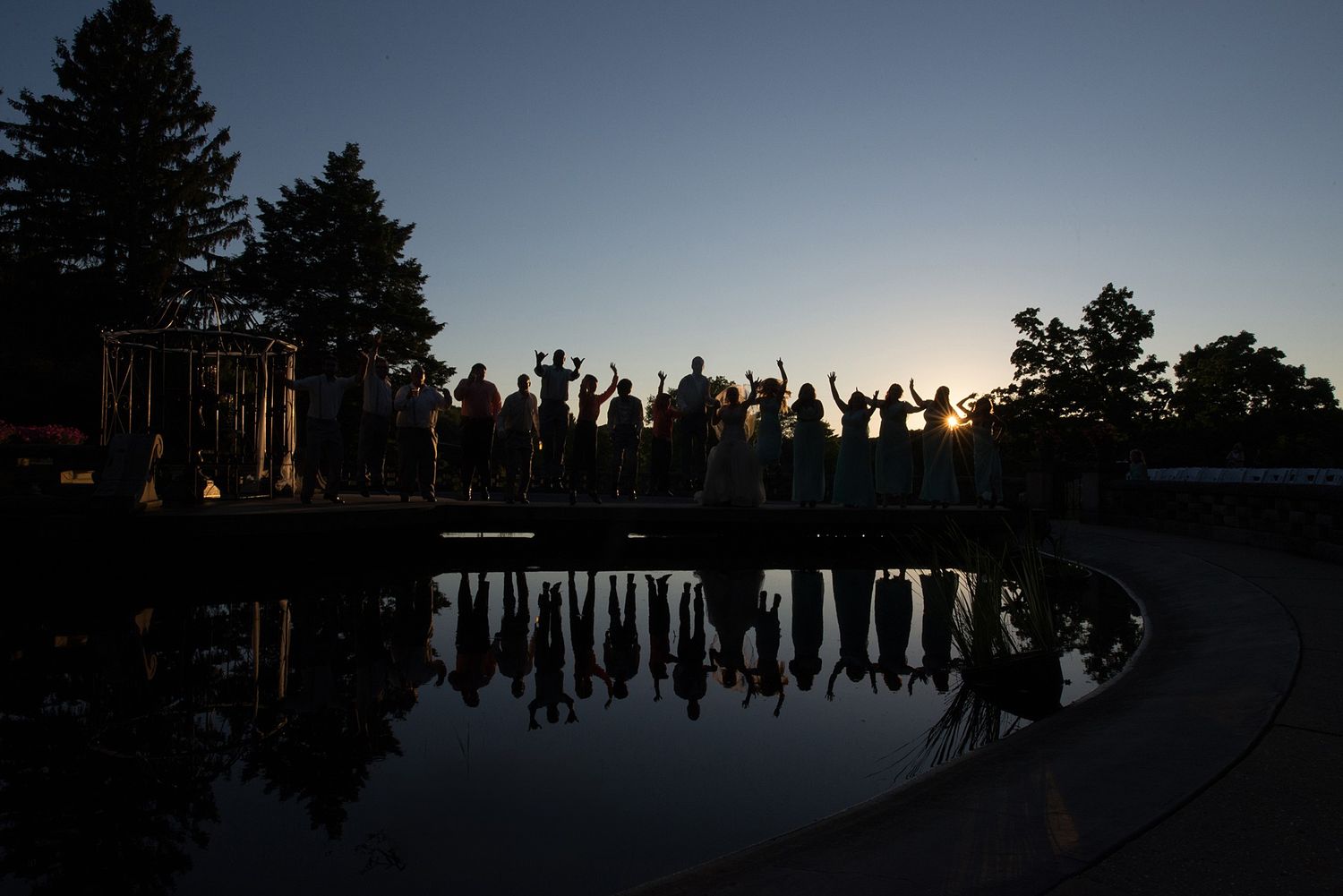 Silhouettes of wedding guests with raised arms reflected in a pool at sunset, surrounded by tall trees.