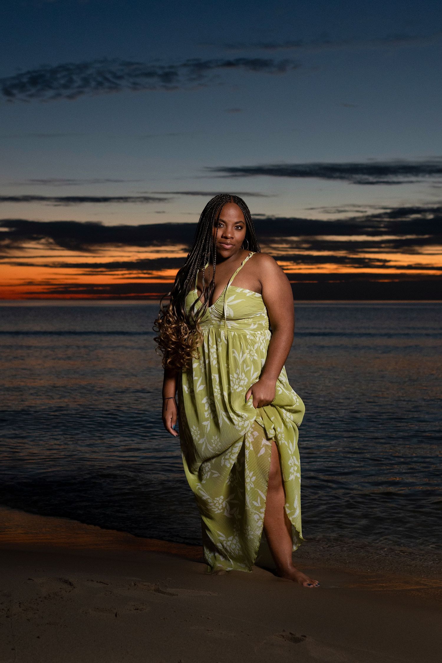Beach photoshoot at dusk with figure in light green dress against dramatic sky.