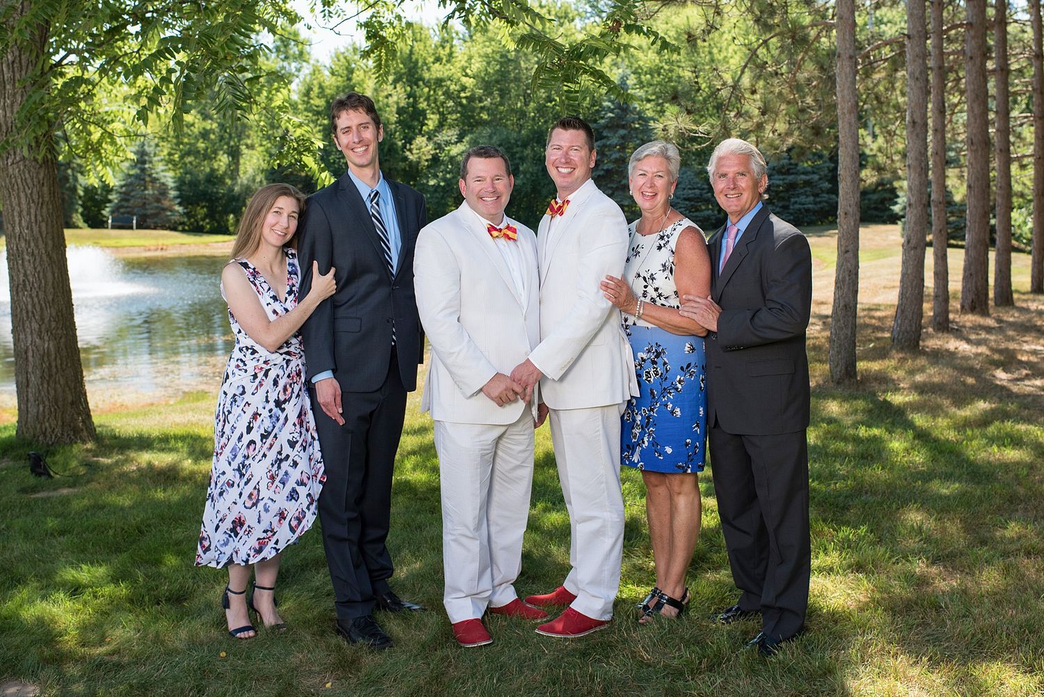 Six people posing outdoors near a pond and trees, dressed in formal attire for a special celebration.