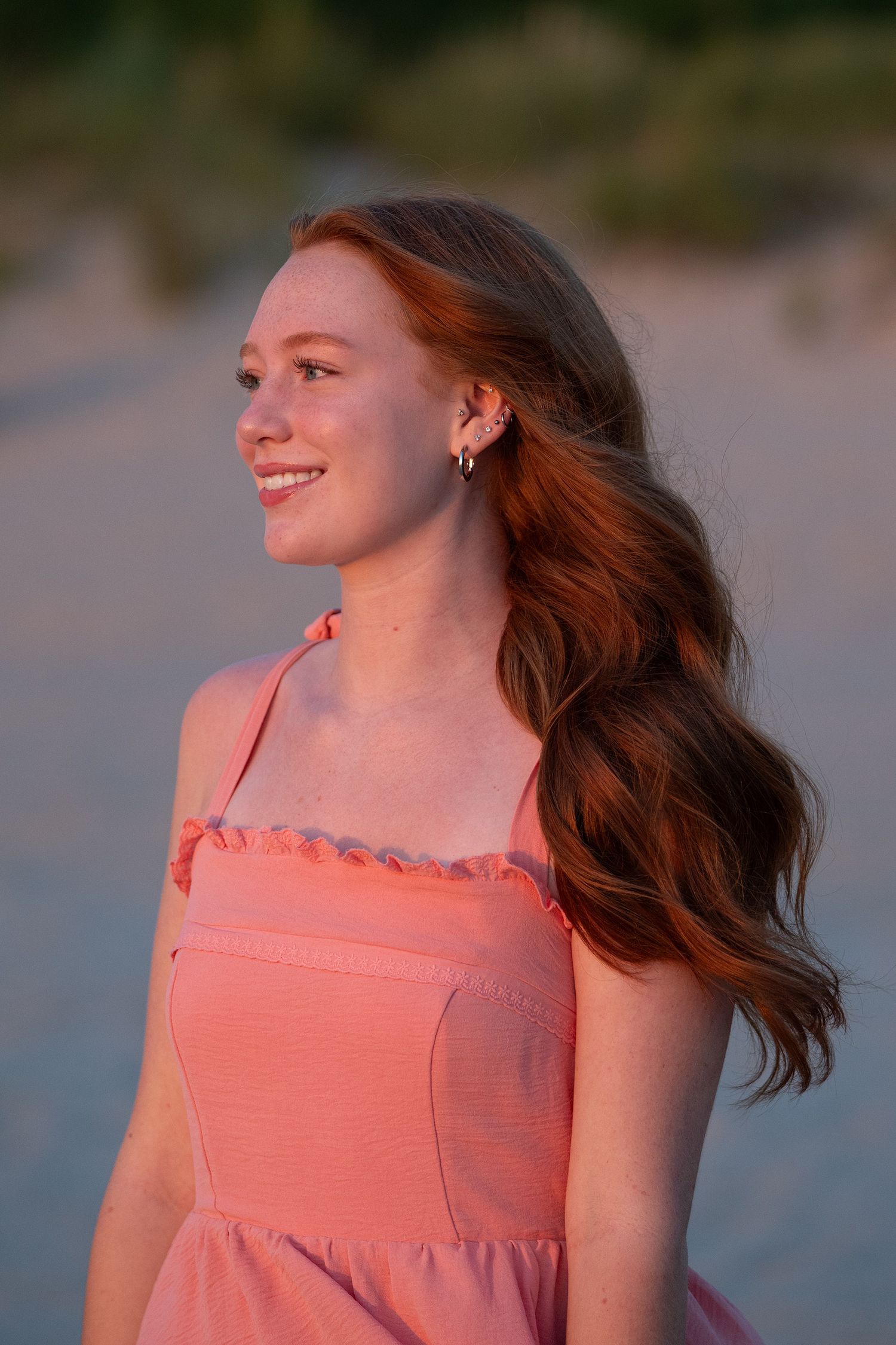 Young woman with long red hair smiling in a pink sundress on a beach at golden hour sunset.