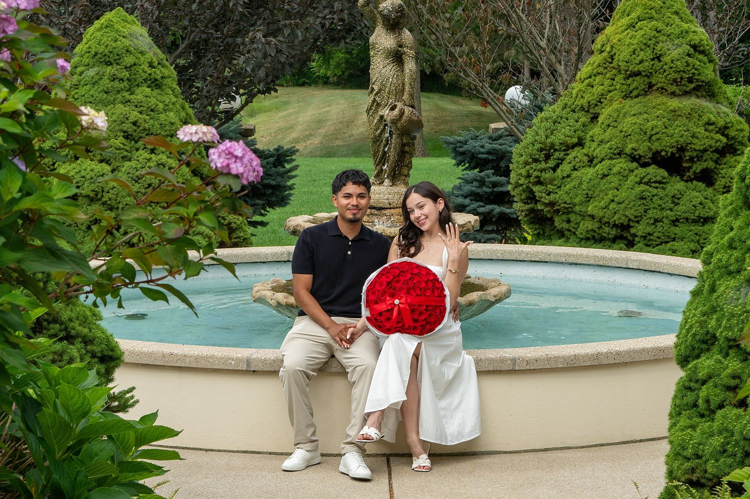 Couple sitting by garden fountain with red rose bouquet, lush greenery and statue backdrop.