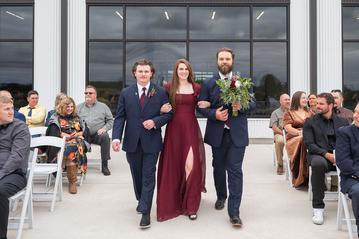 Bridesmaid in burgundy gown walks down wedding aisle escorted by two groomsmen in navy suits.