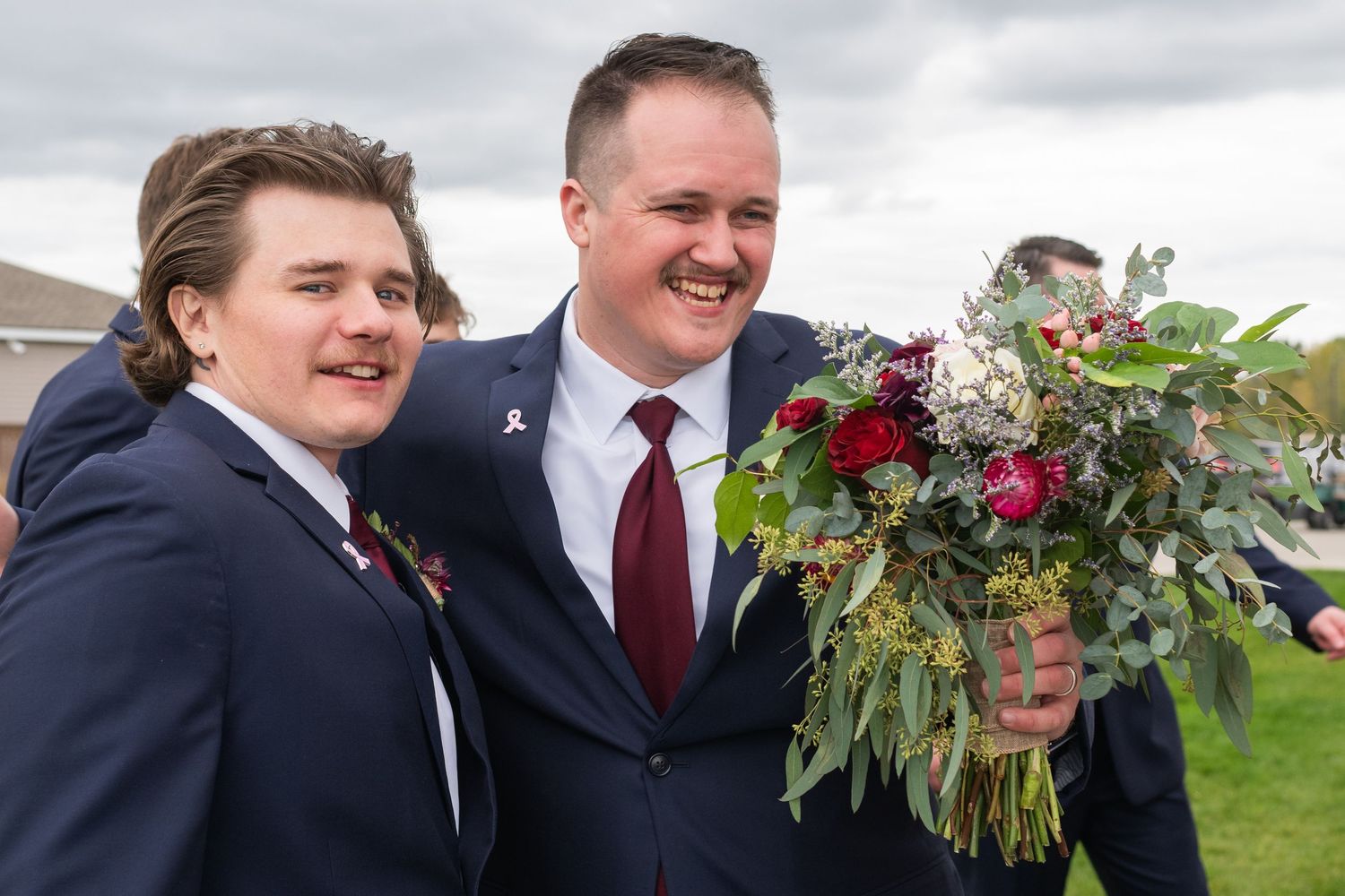 Two men in navy suits smiling outdoors, one holding a bouquet of red roses and greenery at a wedding celebration.
