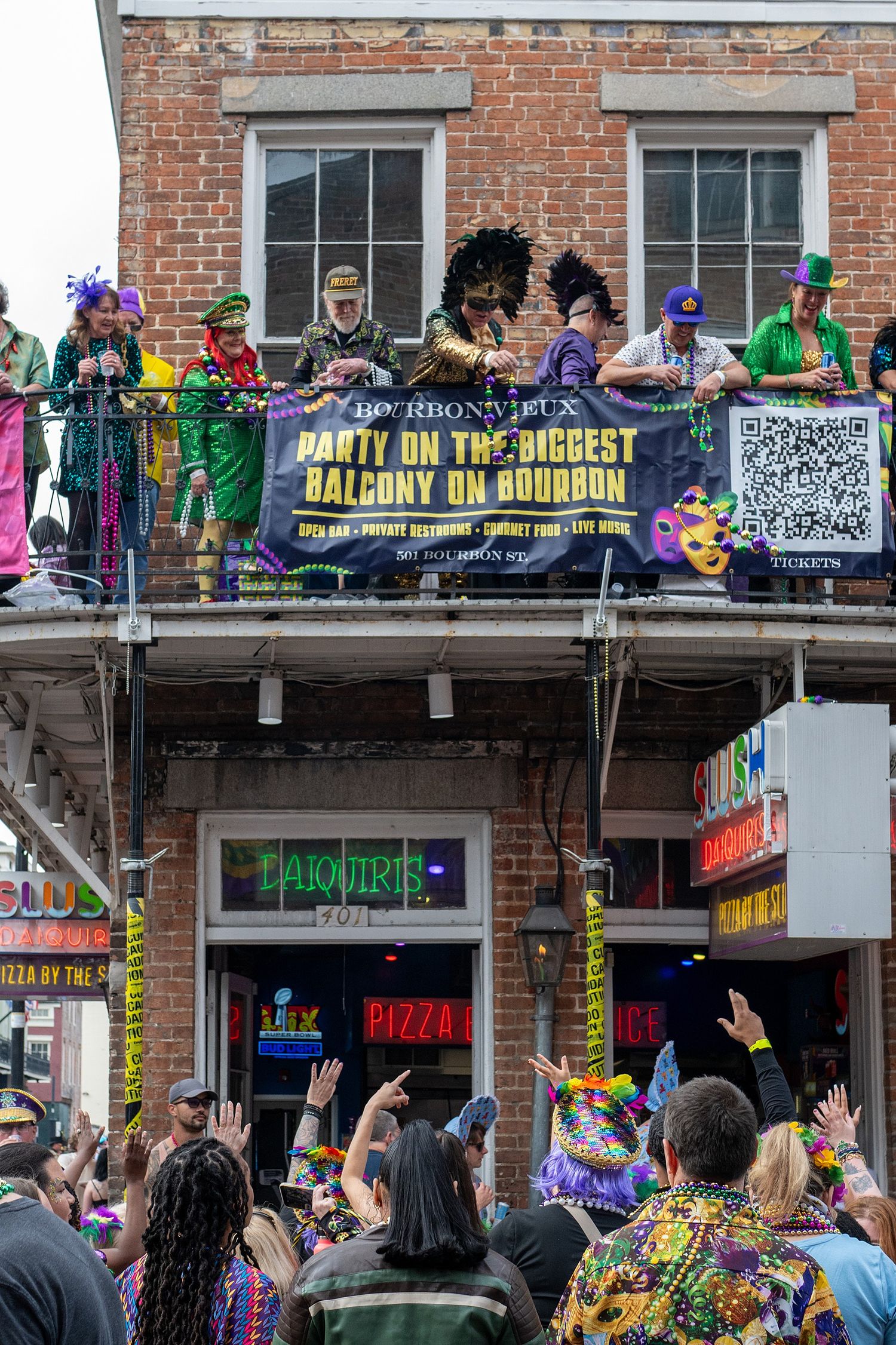 Mardi Gras crowd gathers beneath French Quarter balconies decorated with party banners and neon lights.