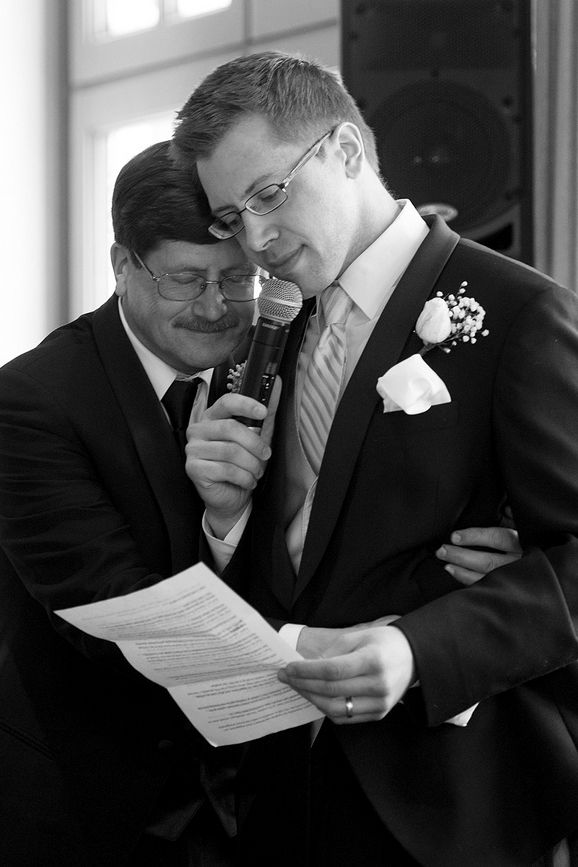Two people share a microphone while reading from papers during a formal event with one wearing a boutonniere.