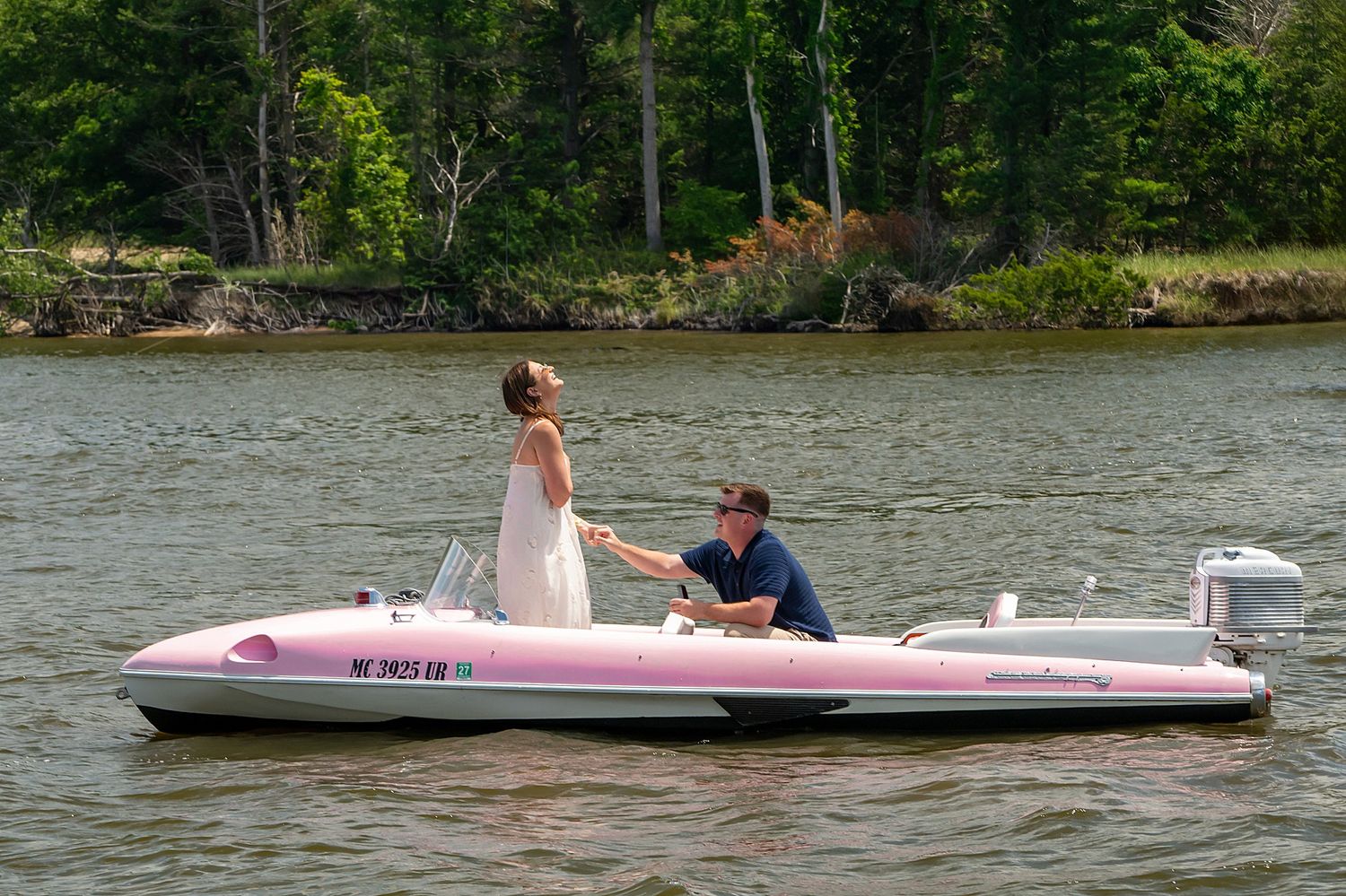 Bride and groom share a romantic moment on a pink motorboat on a calm lake surrounded by lush green trees.