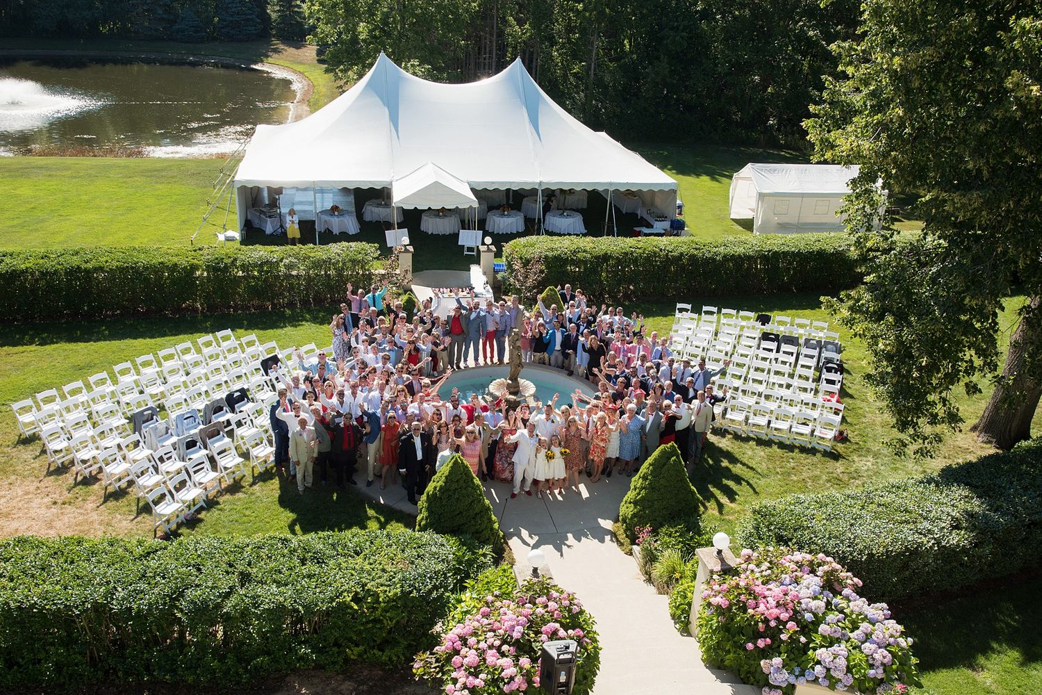 Aerial view of a large outdoor wedding reception with guests gathered on a lawn beside a white tent and floral arrangements.