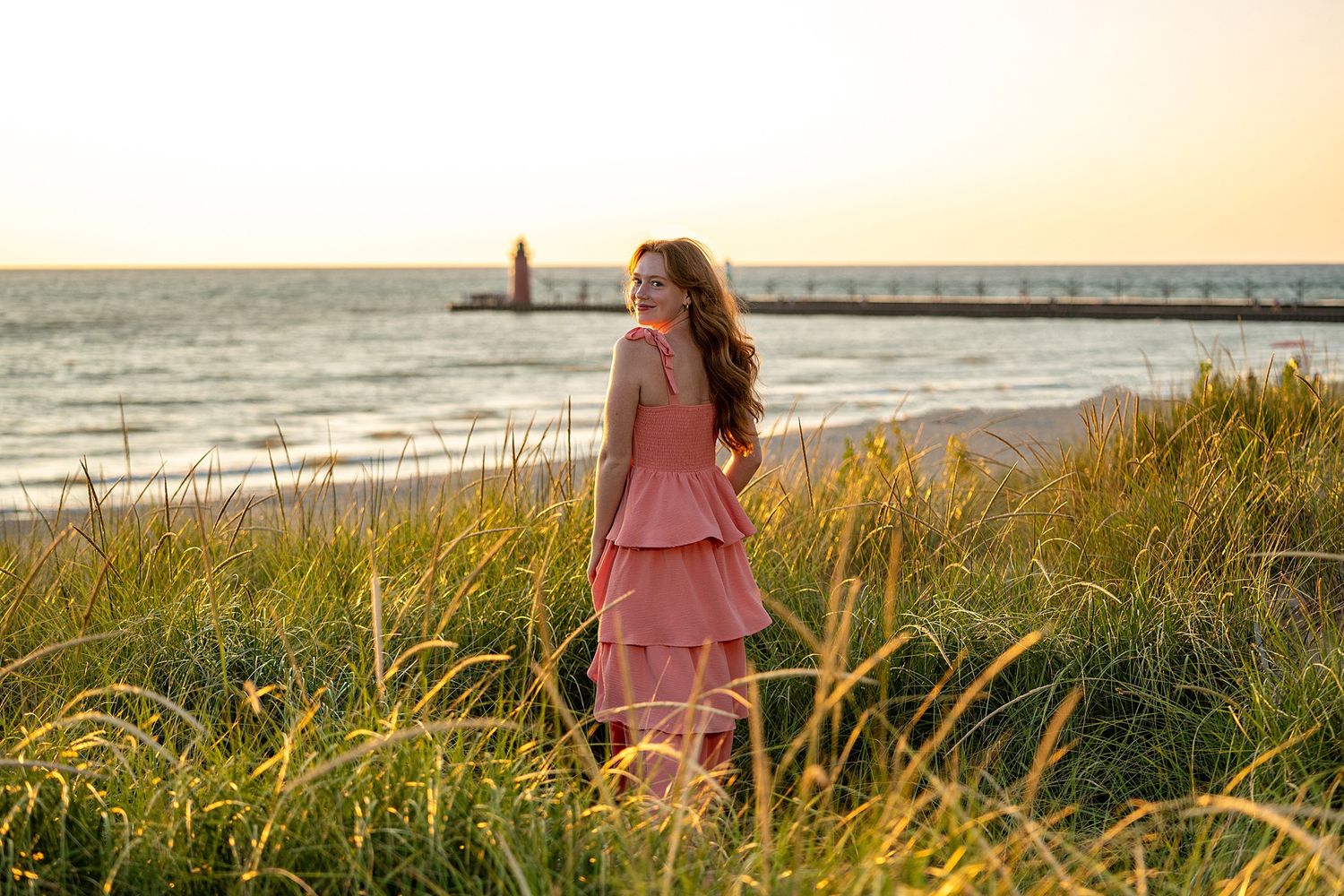 Woman in pink tiered dress standing in beach grass at sunset with lighthouse and ocean in background.