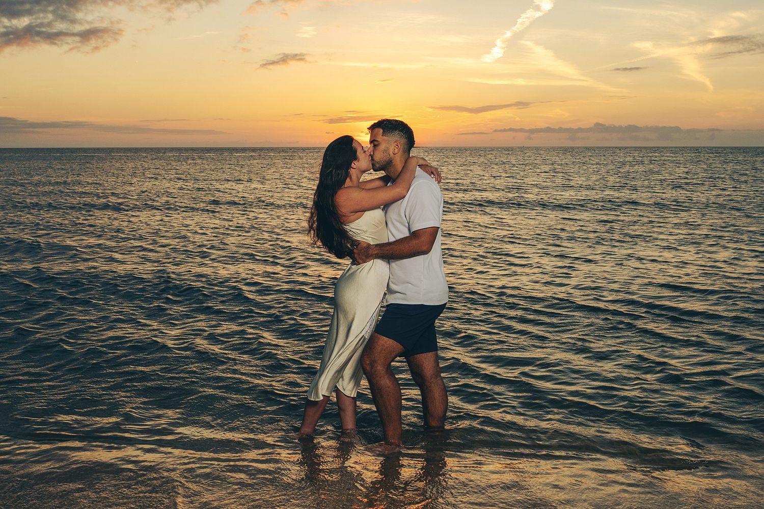 Couple kissing and embracing in shallow ocean water at sunset, with golden skies and calm waves in the background.