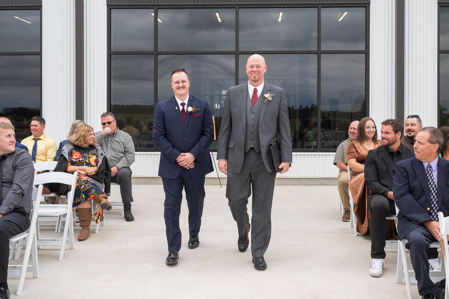 Two men in suits walk down outdoor wedding ceremony aisle together.