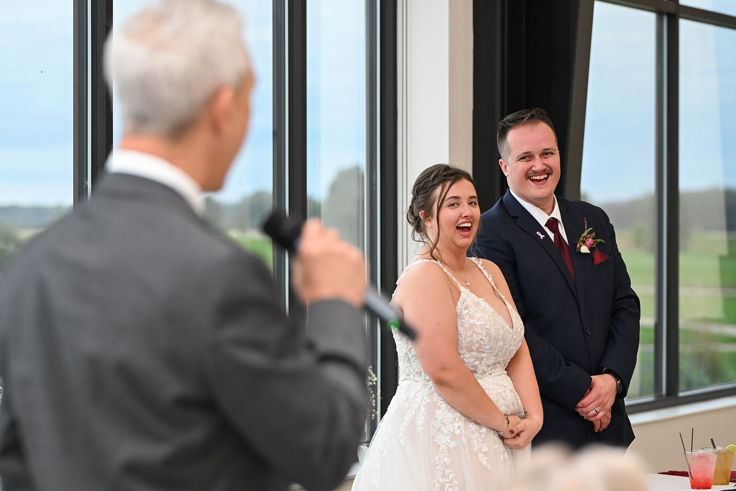 Bride and groom laughing joyfully during a wedding toast speech at a modern venue with large windows.
