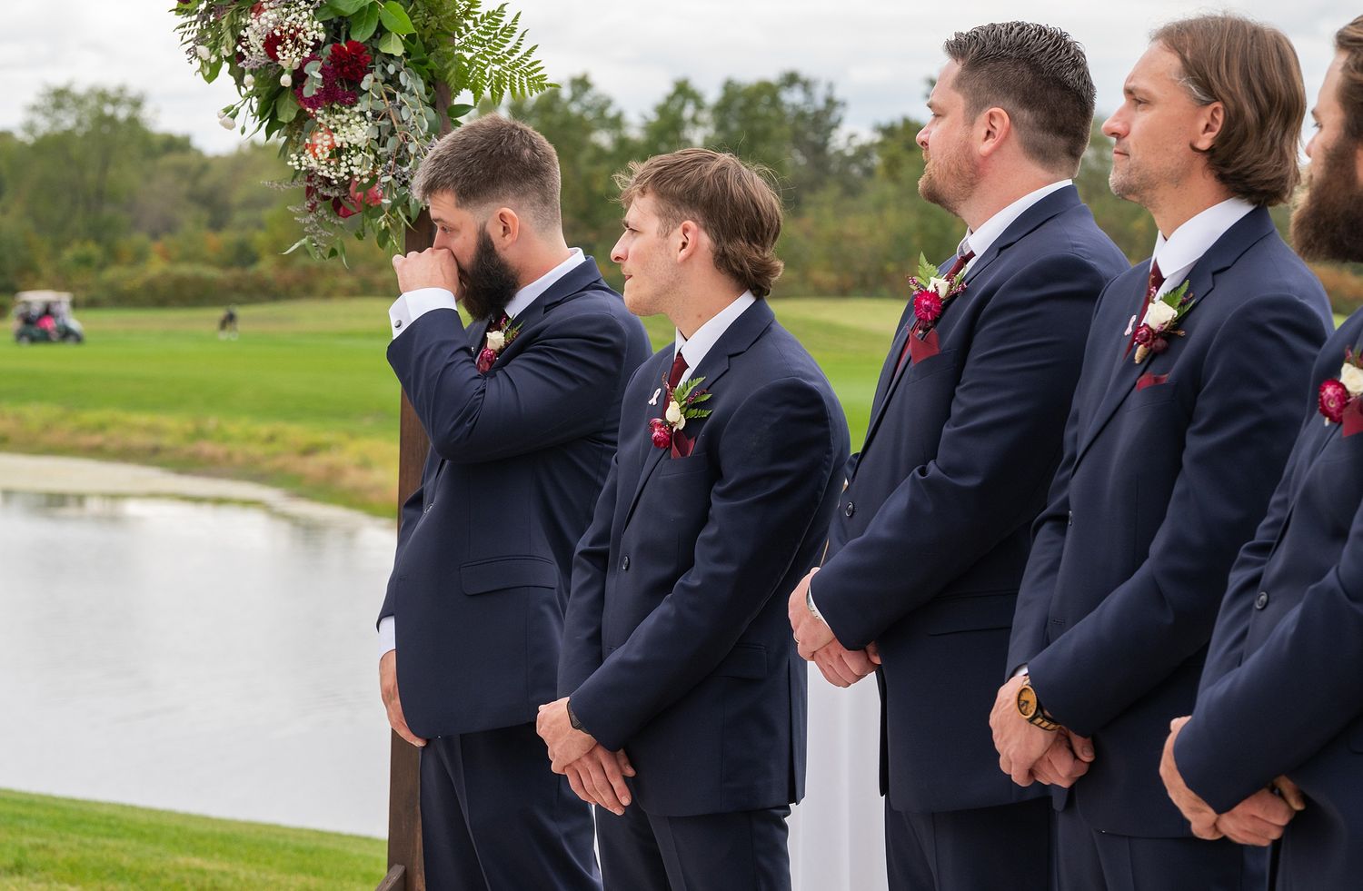 Groomsmen in navy suits stand at outdoor wedding ceremony beside floral arch near pond.