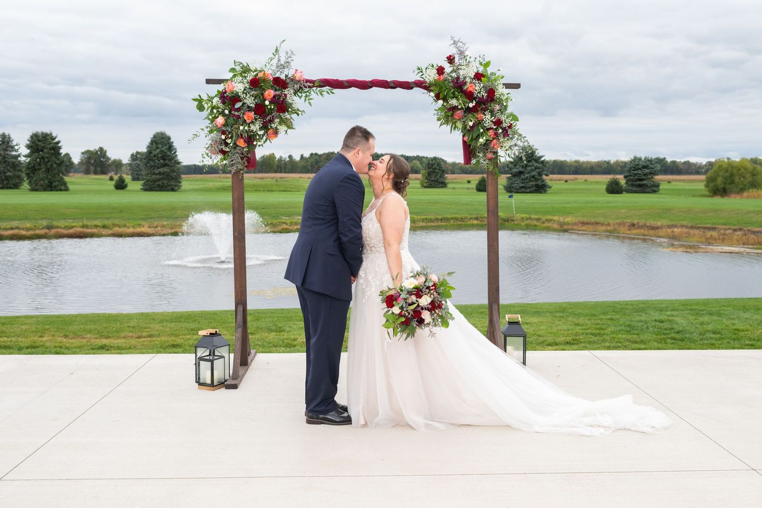 Bride and groom share first kiss under floral arch at outdoor wedding ceremony by a pond.