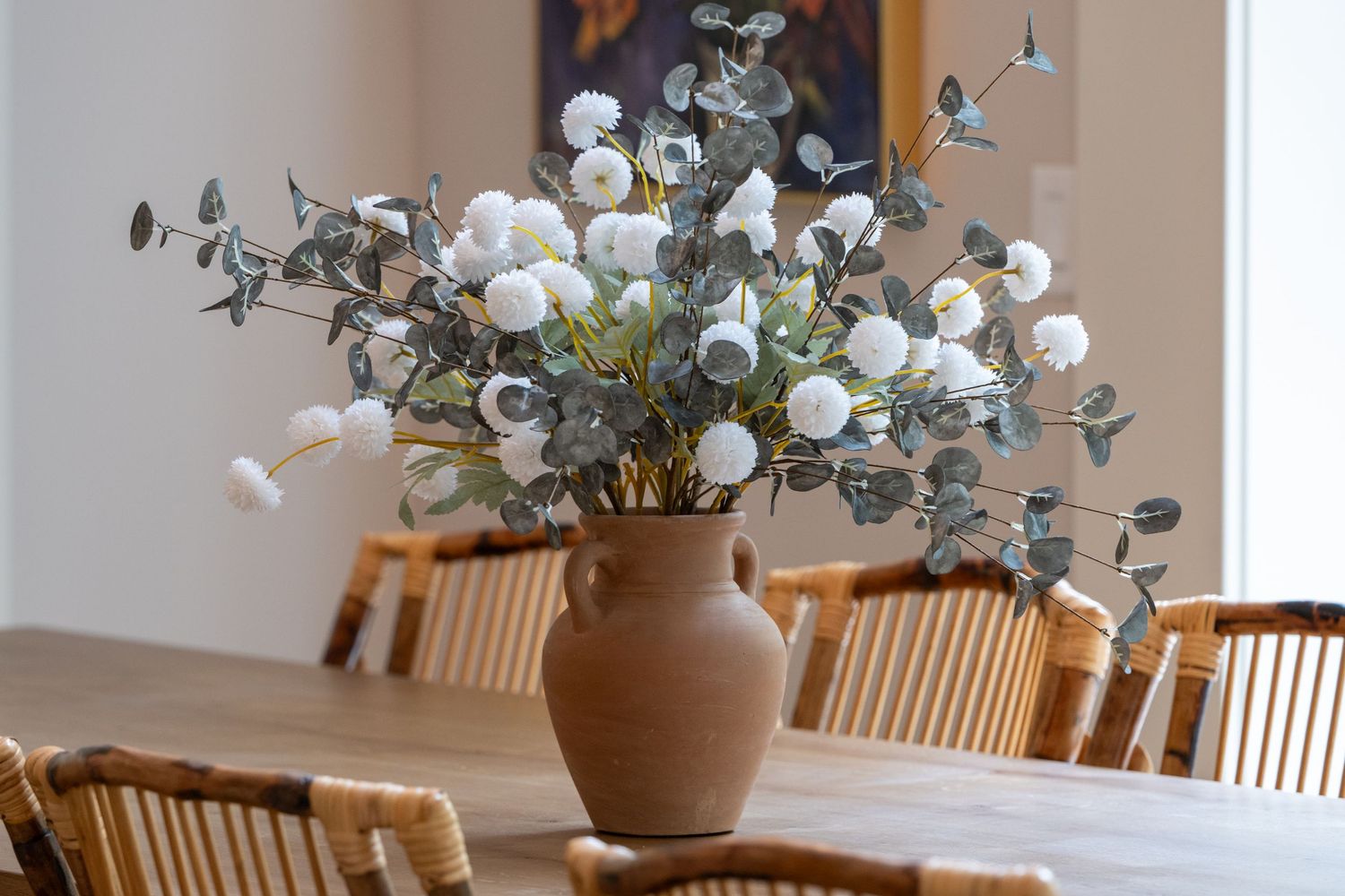 Terracotta vase with white billy balls and eucalyptus on a wooden dining table with rattan chairs.