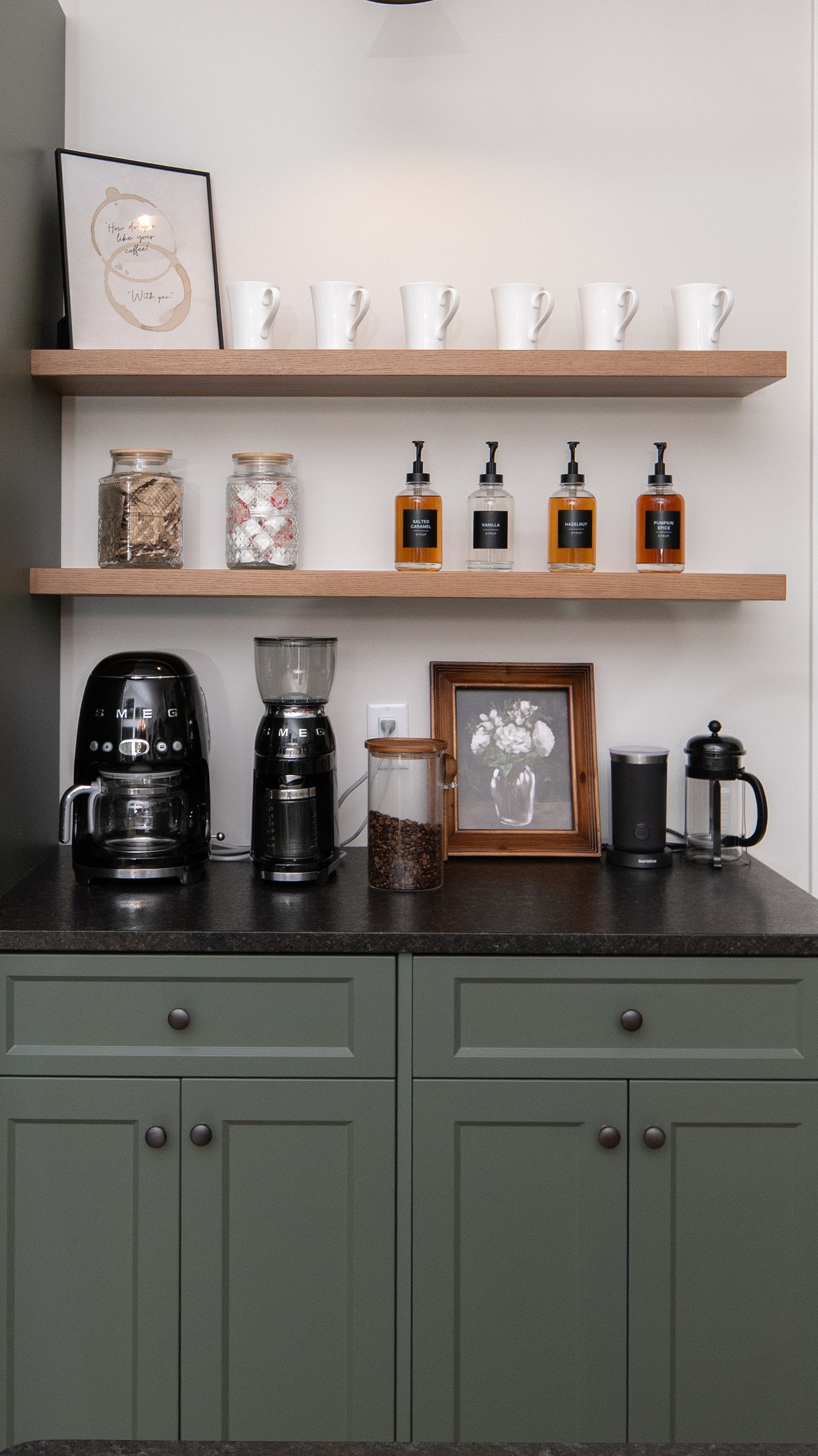 Stylish coffee bar with green cabinets, floating wood shelves, SMEG coffee maker, French press, mugs, and amber syrup bottles.