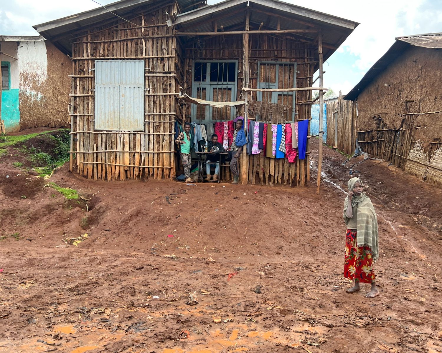 Simple wooden houses with tin roofs stand along a muddy village path.