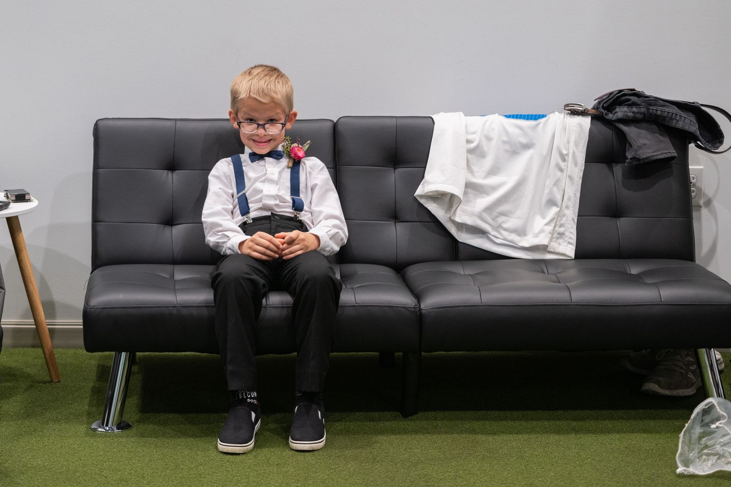 Young ring bearer in suspenders and bow tie sitting alone on a black leather couch before the wedding ceremony.