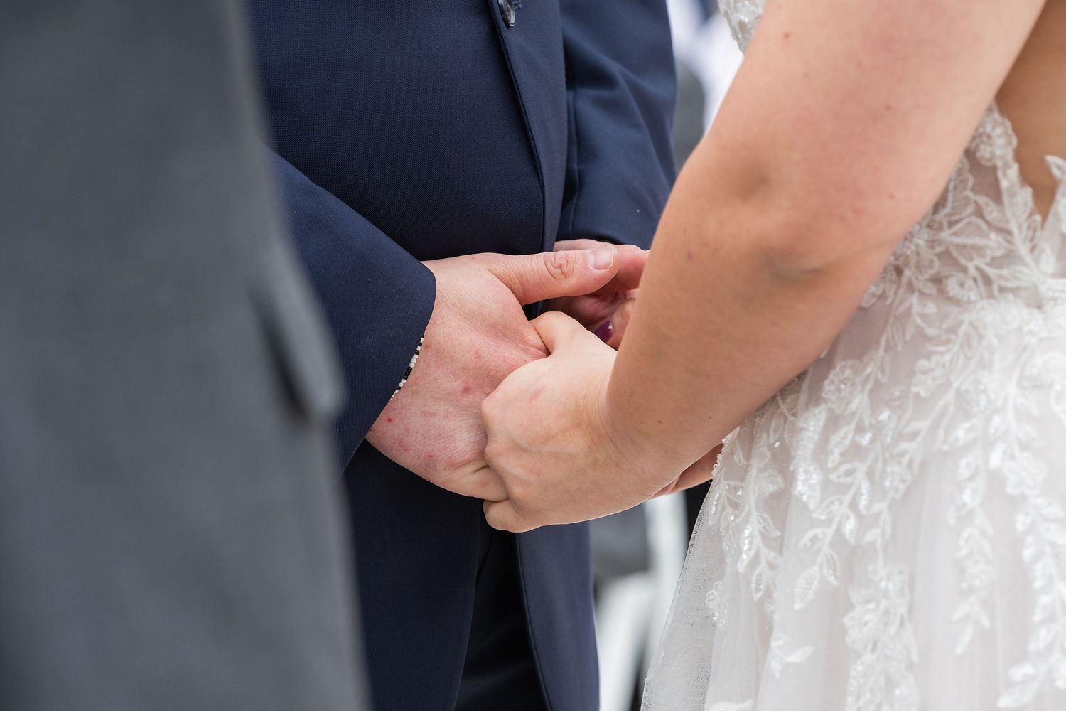 Bride and groom hold hands tenderly during their outdoor wedding ceremony, bride wearing lace gown with pearl bracelet.