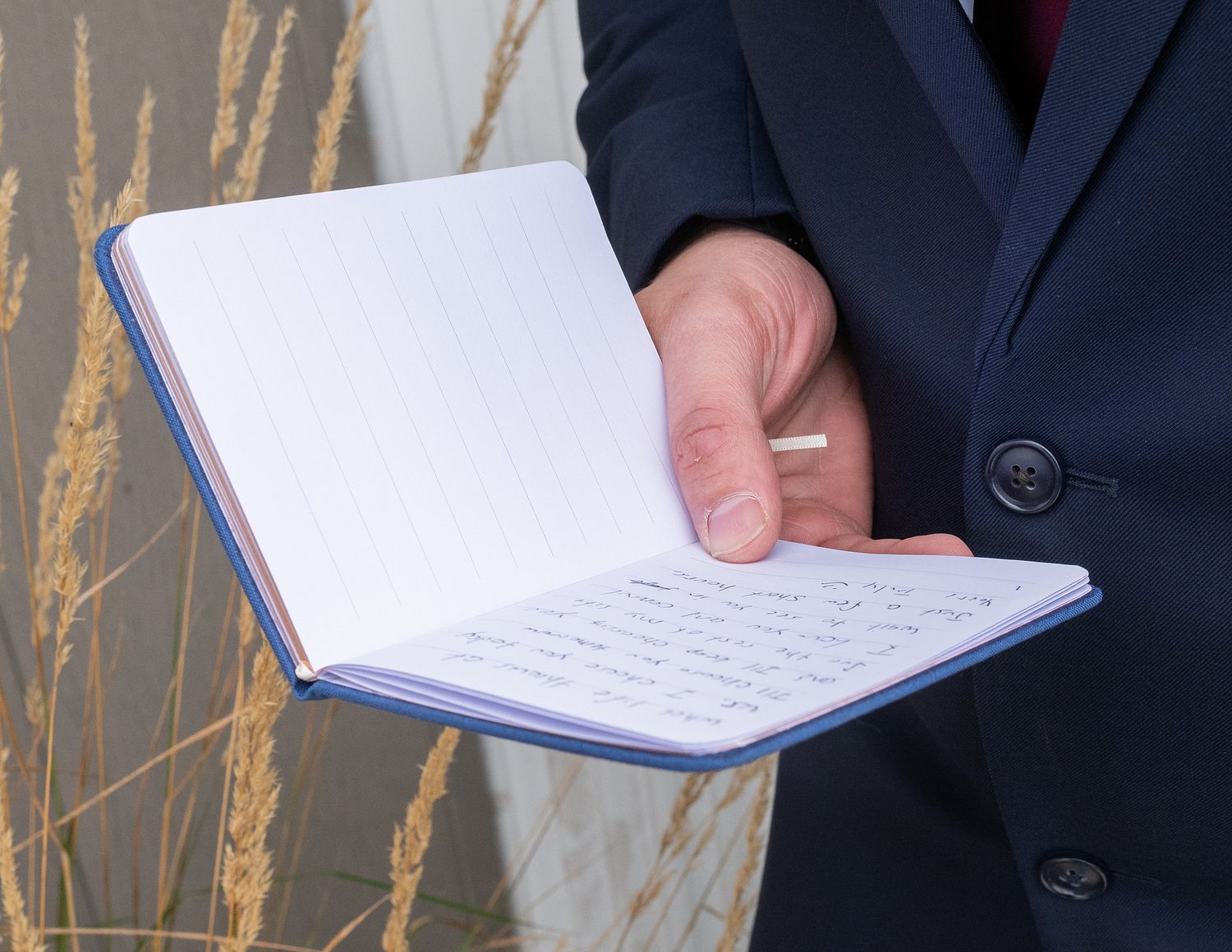 Groom in navy suit holds open a small notebook with handwritten wedding vows during outdoor ceremony.
