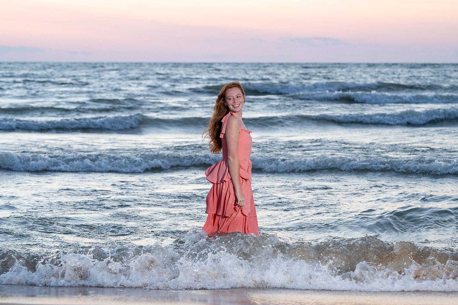 Young woman in pink dress standing in ocean waves at sunset on a sandy beach.