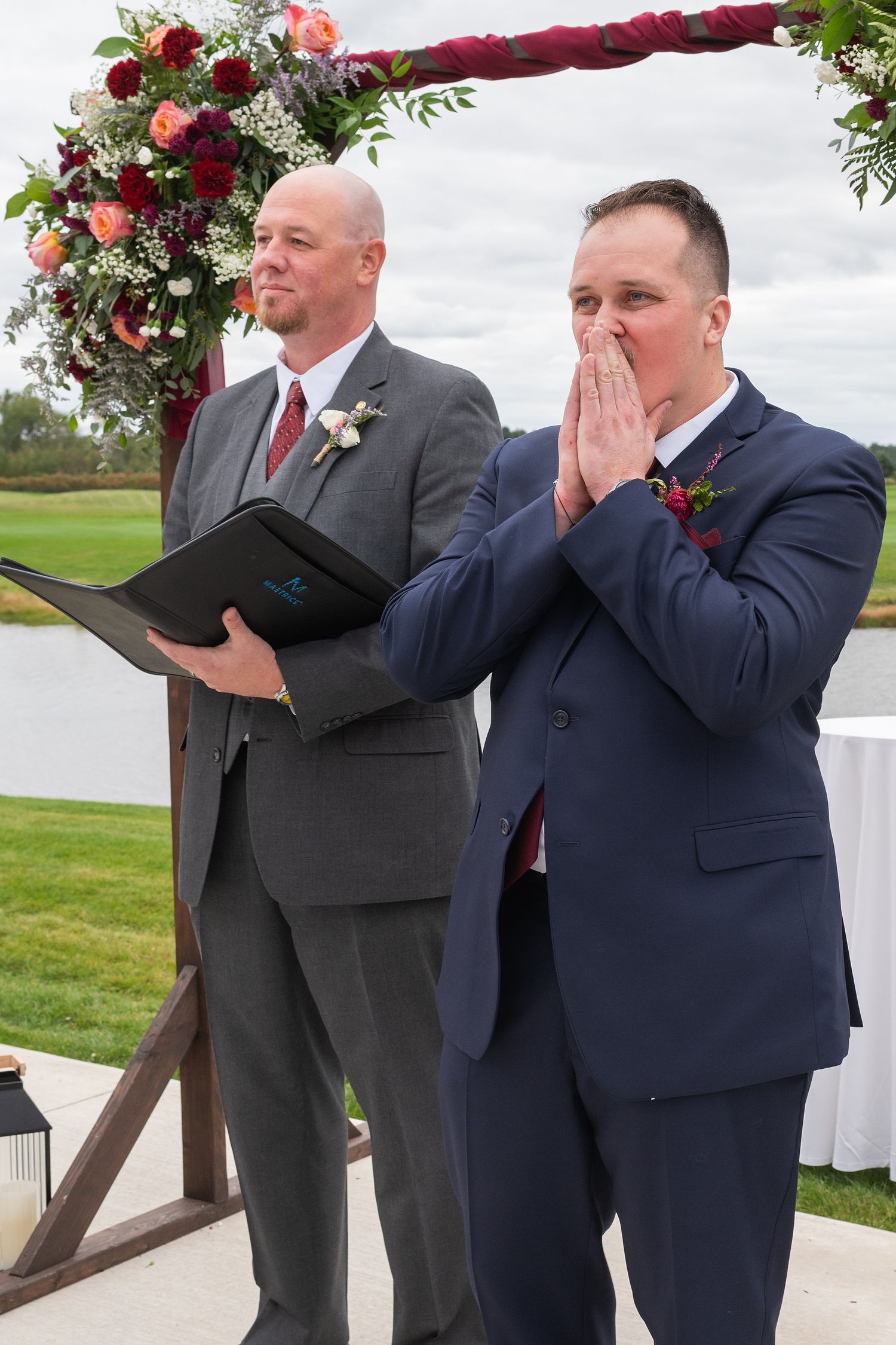 Groom in navy suit clasps hands emotionally at outdoor wedding altar.