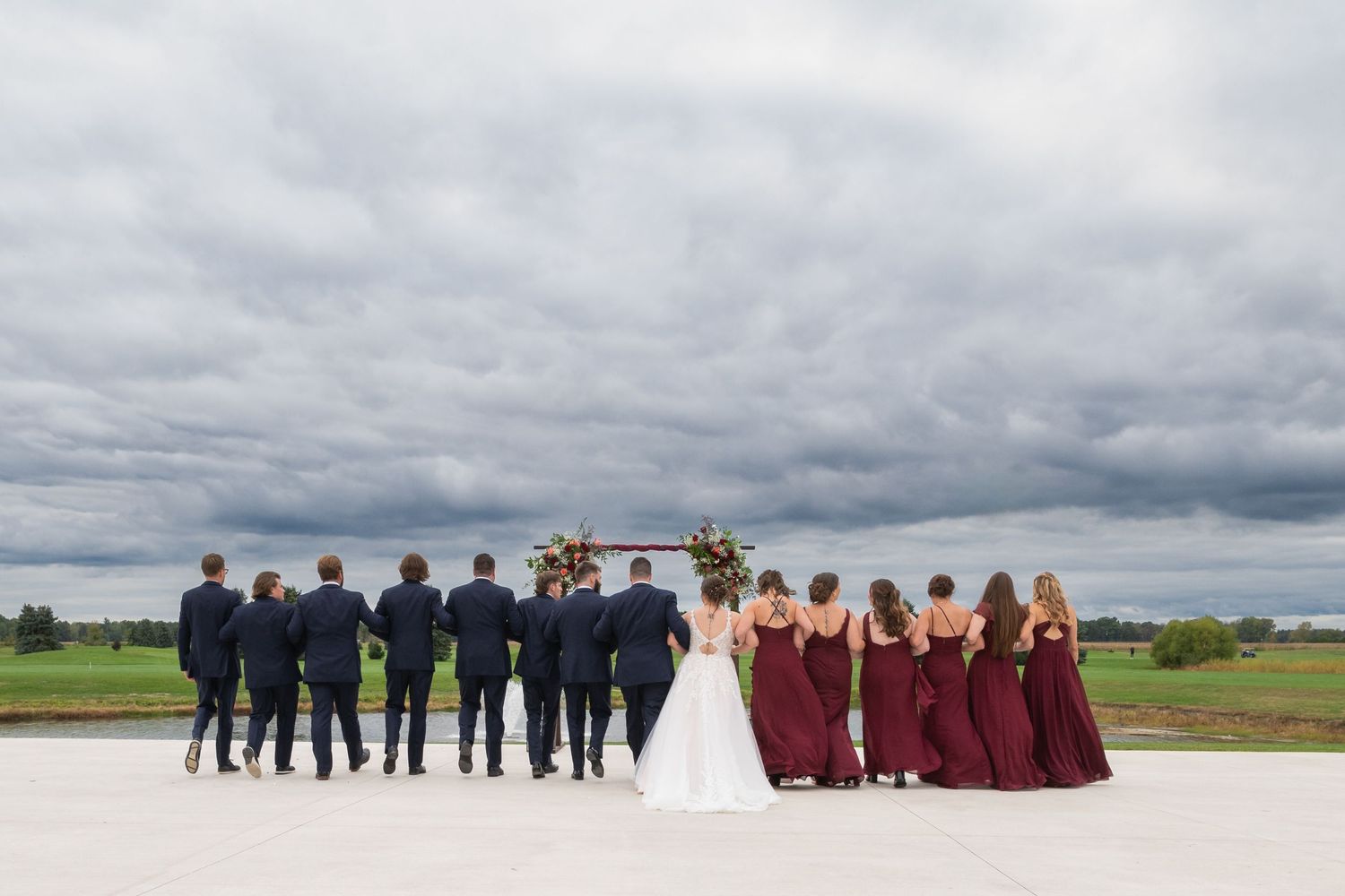Wedding party in navy suits and burgundy gowns stands before a floral arch under dramatic cloudy skies.