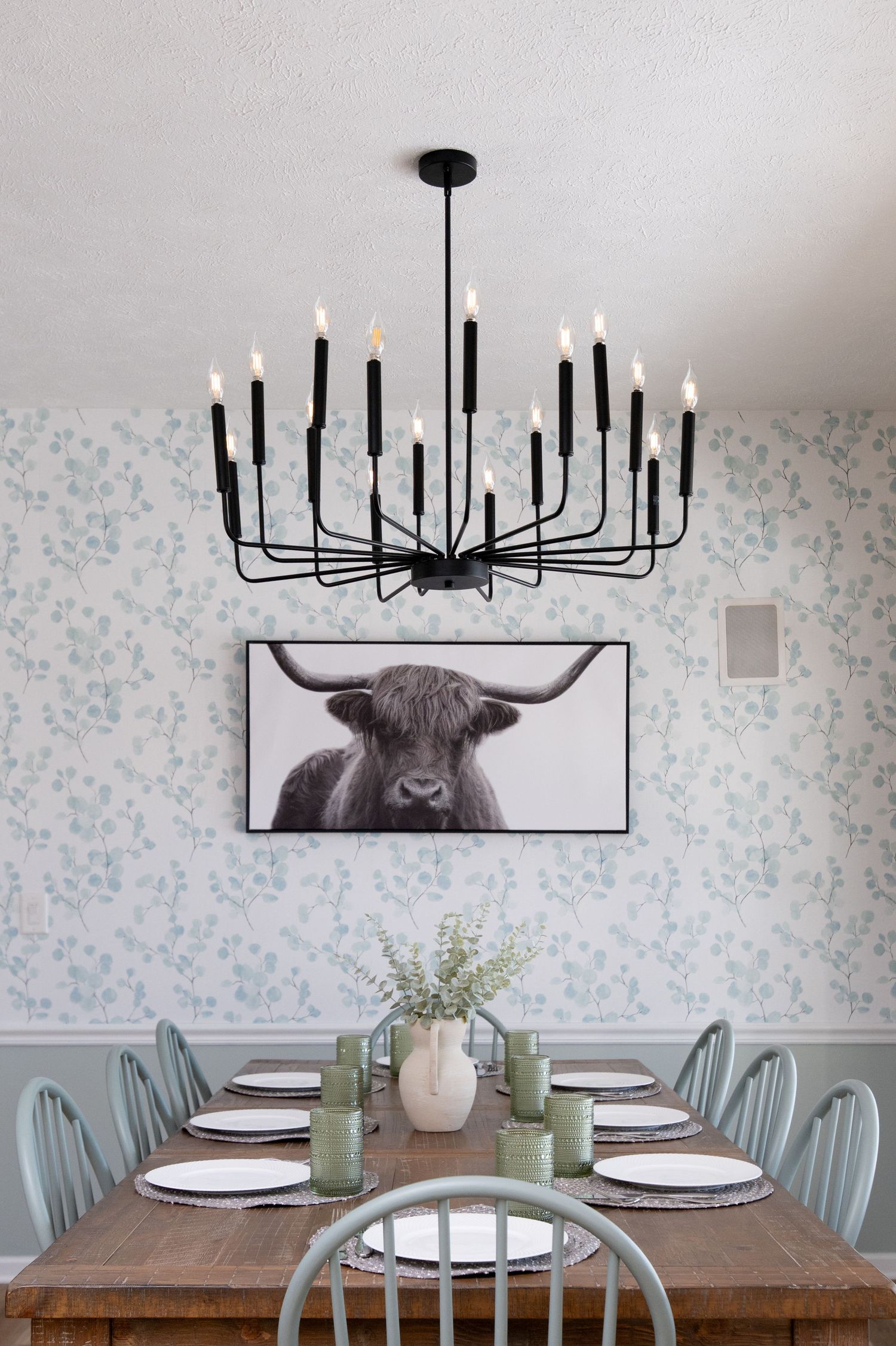 Dining room with black candelabra chandelier, floral wallpaper, farmhouse table, sage chairs, and highland cow wall art.