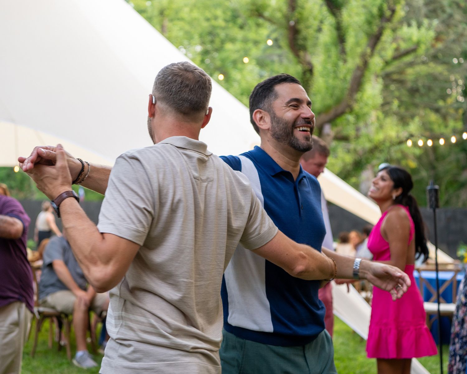 People dancing and celebrating at an outdoor party with tents and string lights visible in the background.