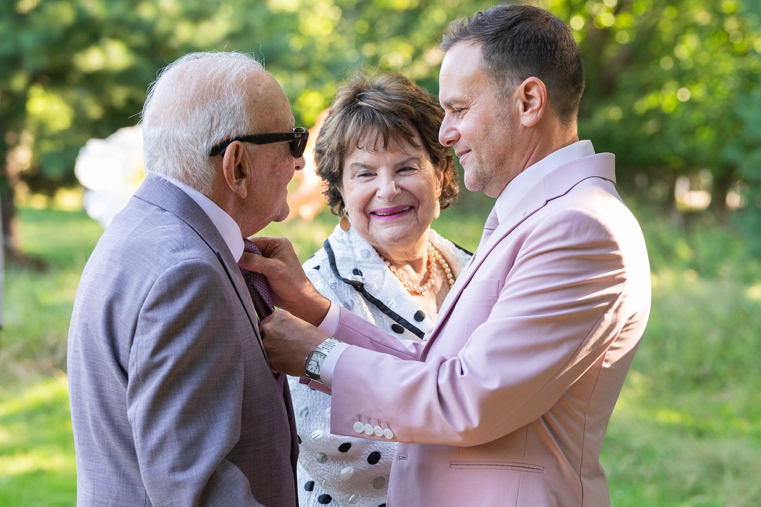 Three people share a warm moment together outdoors on a sunny day in a green park setting.