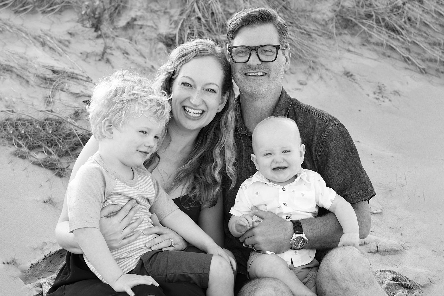 A black and white beach portrait of a family sitting together in the sand.