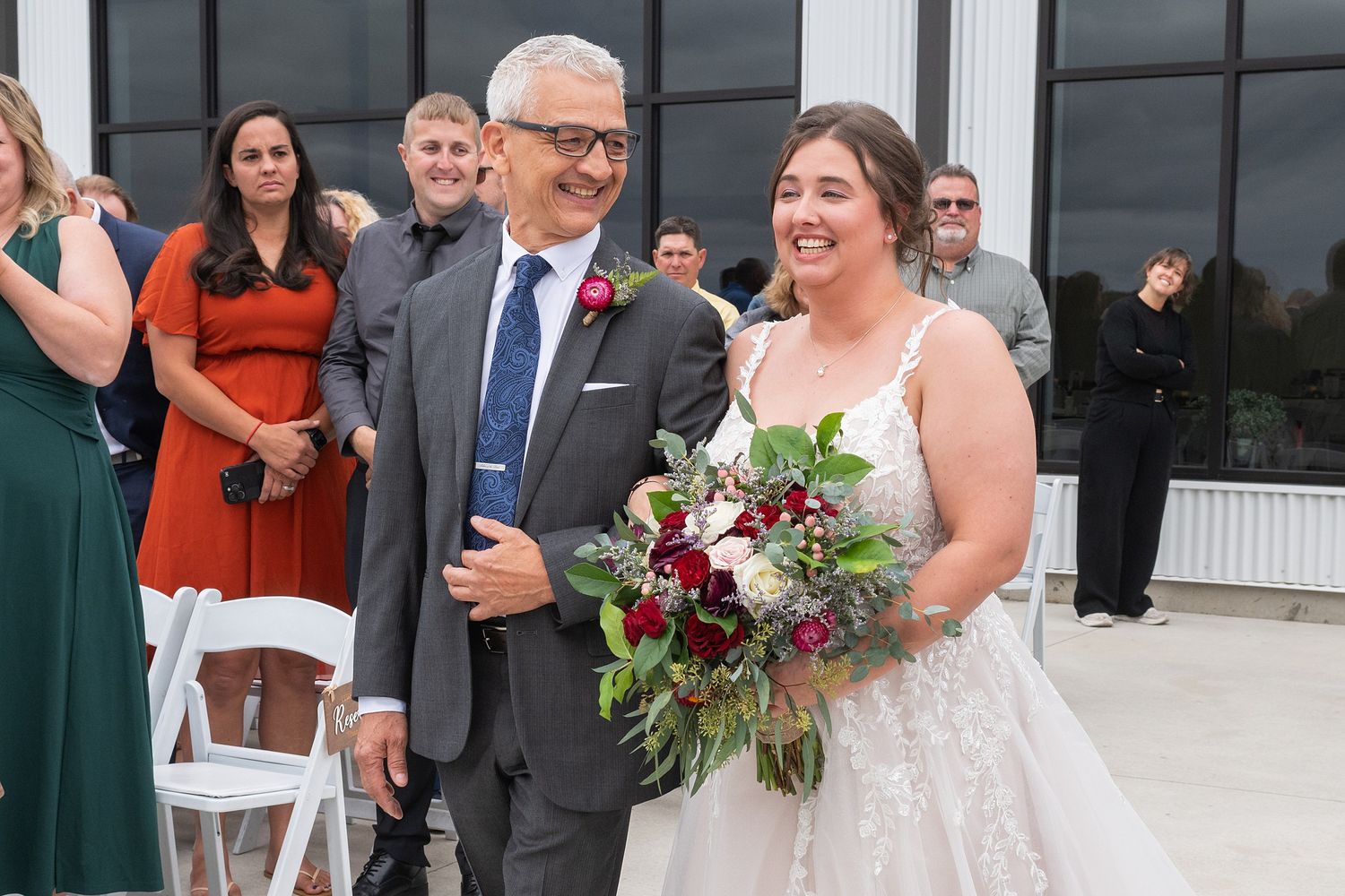 Father walks bride down the aisle at outdoor wedding ceremony, bride holding lush burgundy bouquet.