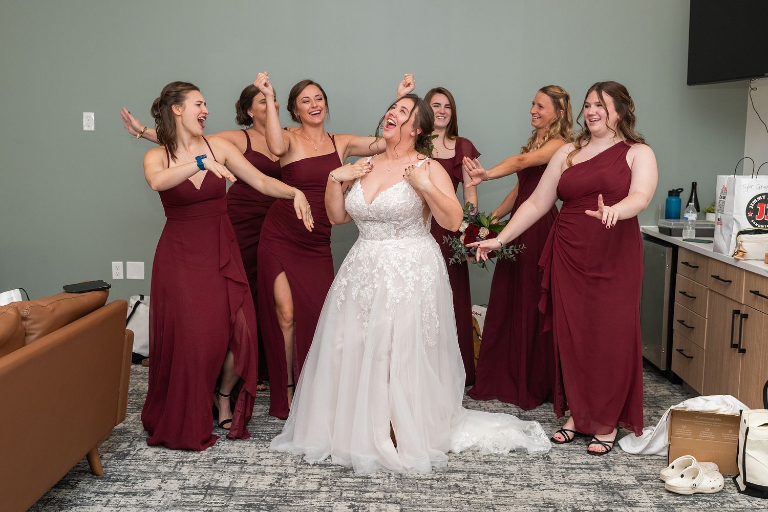 Bride in white gown surrounded by bridesmaids in burgundy dresses celebrating joyfully in a bridal suite.